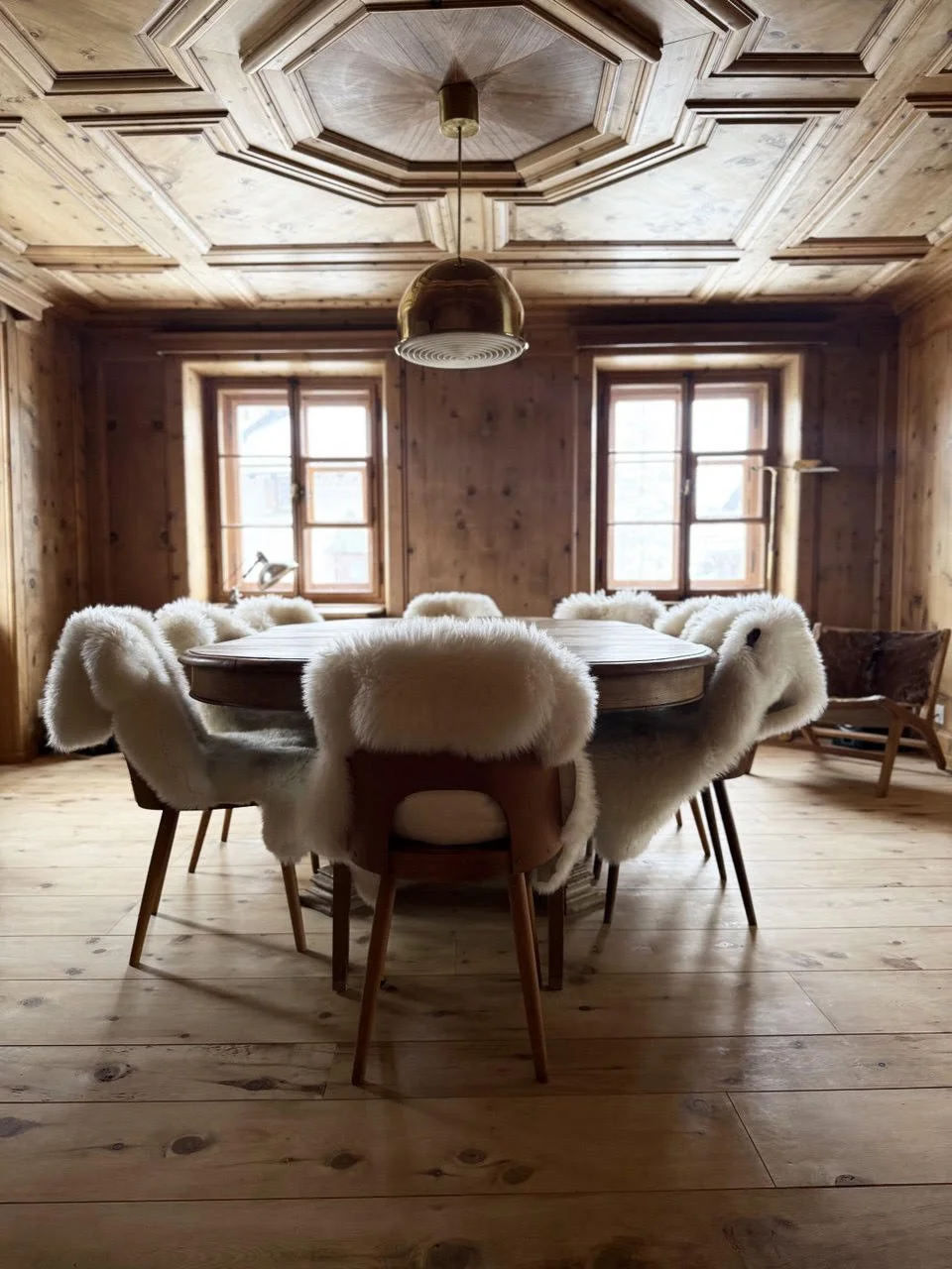 Sala da pranzo in legno con tavolo rotondo e sedie con rivestimenti di pelliccia bianca. Due finestre con cornici di legno e una lampada sospesa di colore oro.