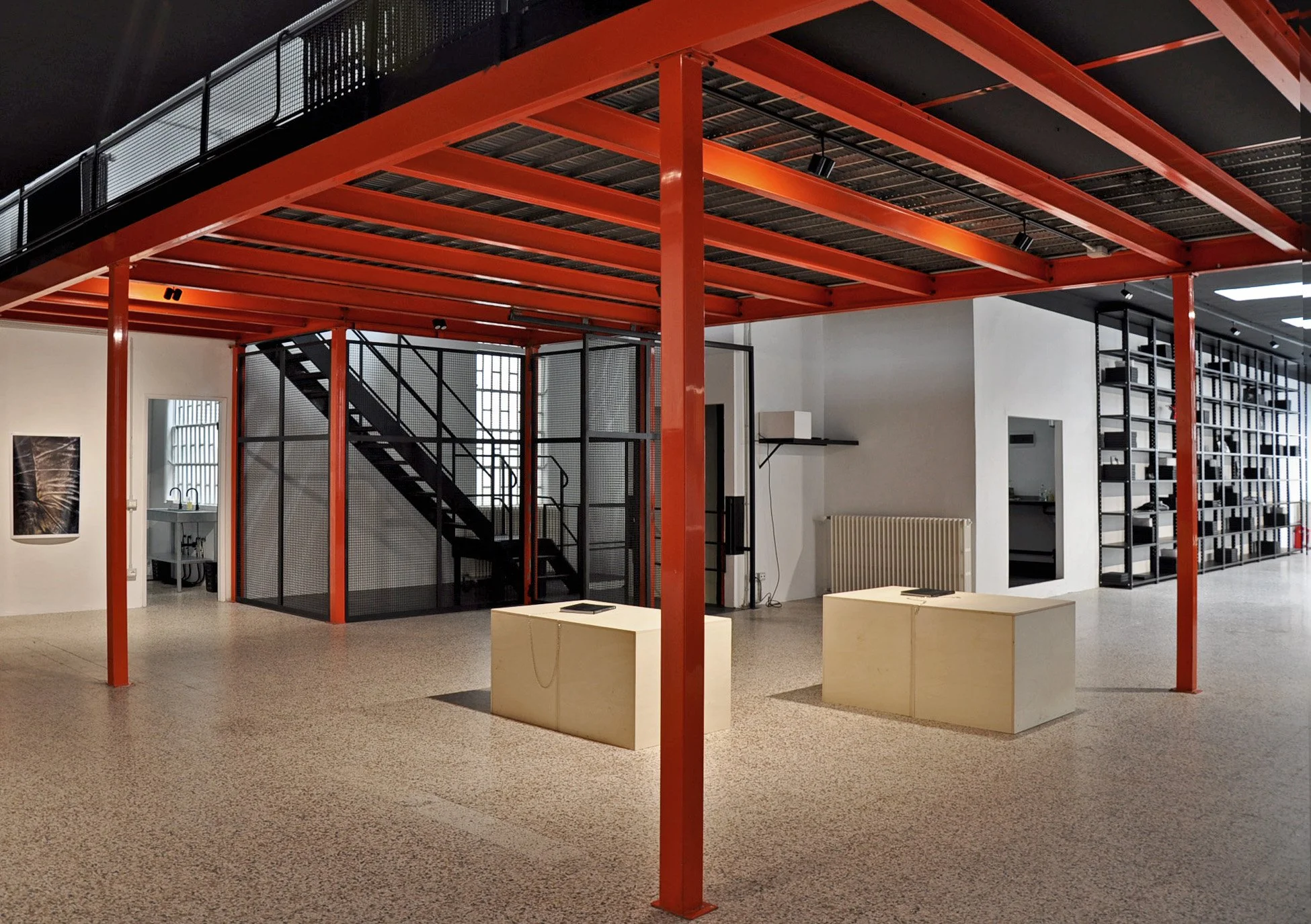 Interno di un museo o centro culturale con strutture in metallo nero e rosso, aree espositive e scaffalature.