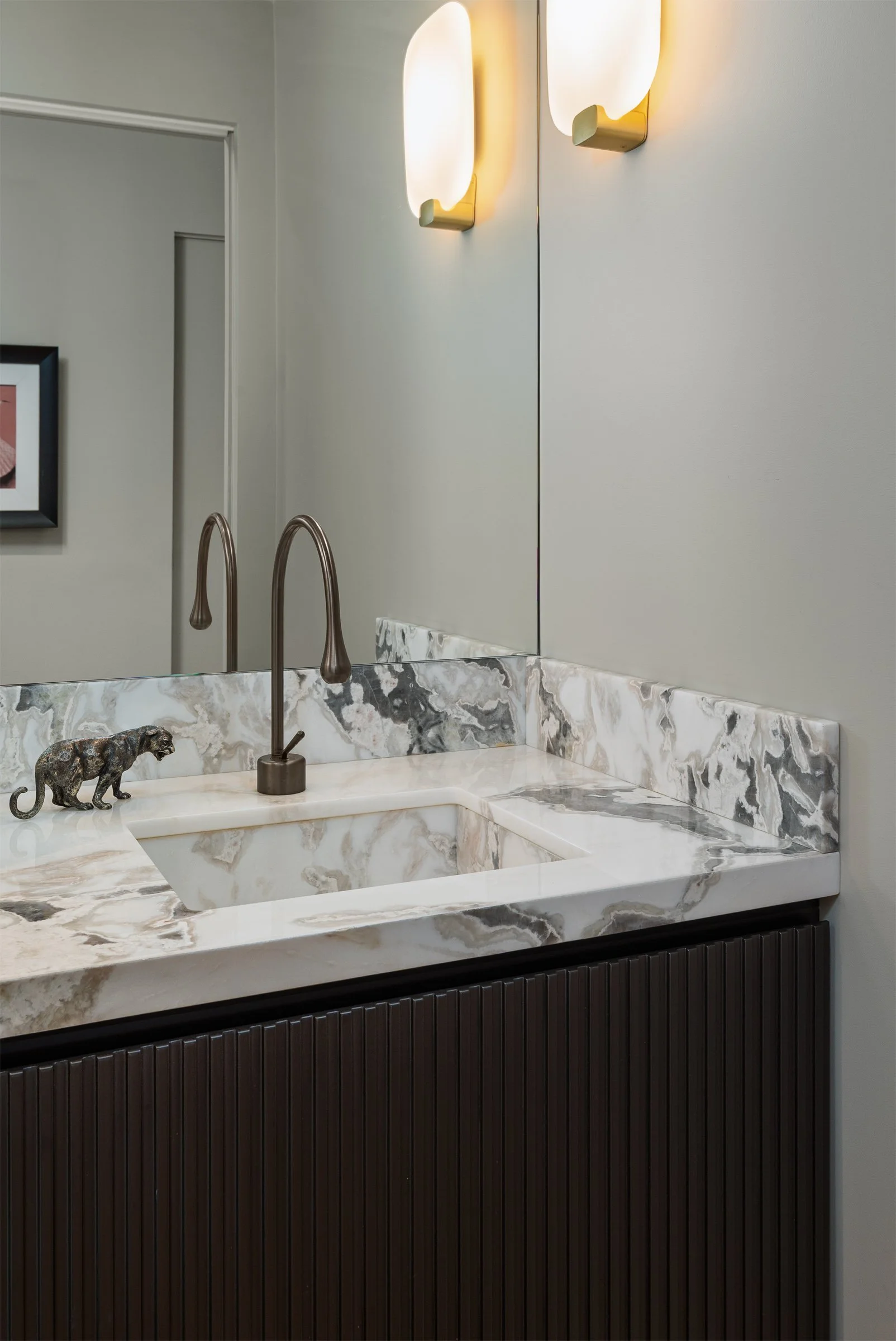 Lavabo con piano in marmo con venature beige, bianco e grigio, con un rubinetto in ottone. Sopra il lavabo, uno specchio grande e luci armoniche, e sulla sinistra un quadro appeso alla parete.