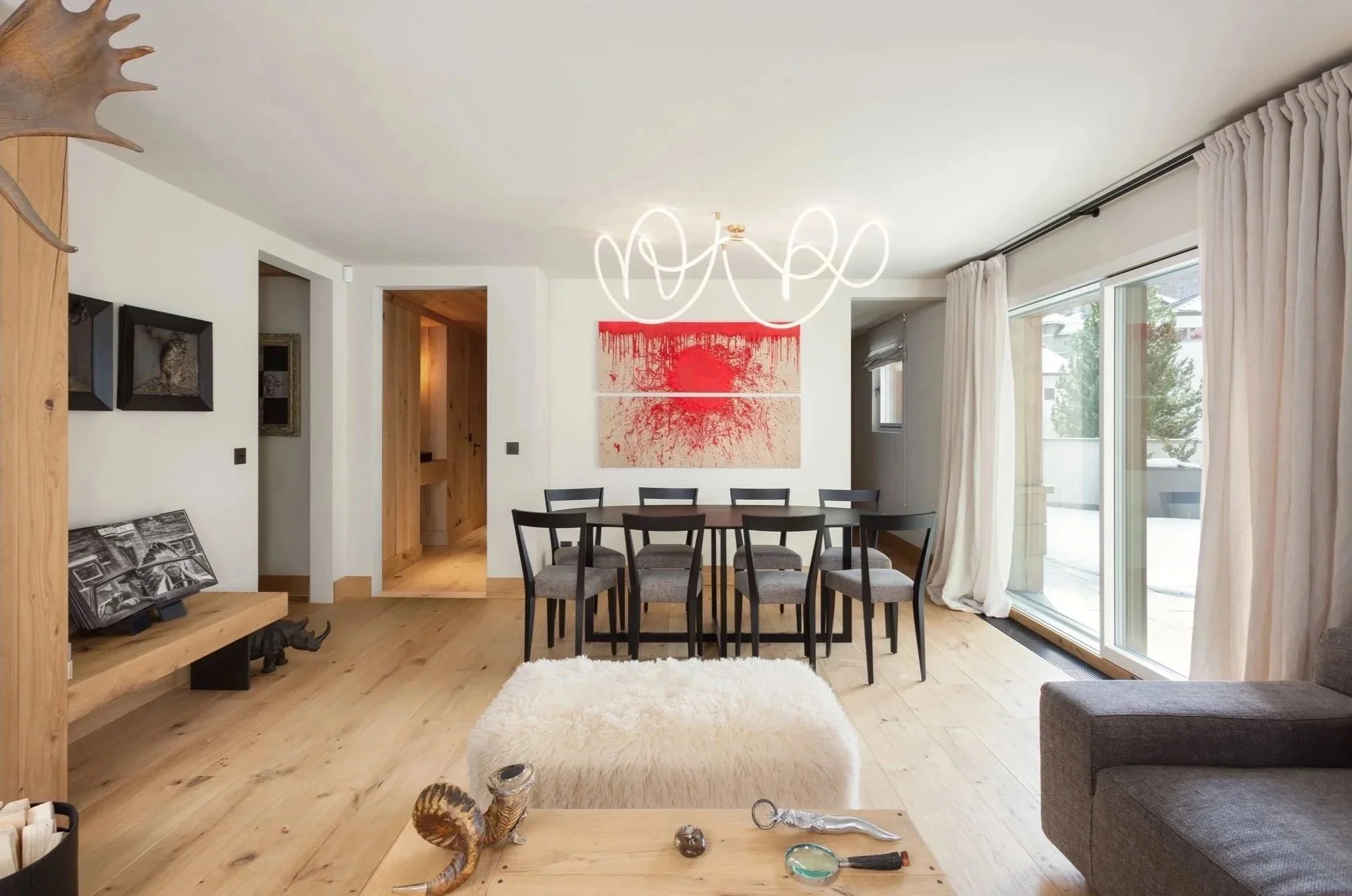 Interno di una sala da pranzo moderna con tavolo e sedie nere, grande finestra con tende bianche, pareti bianche, quadro rosso sopra il tavolo, lampadario luminoso a spirale, pavimento di legno, divano grigio e tappeto bianco soffice.