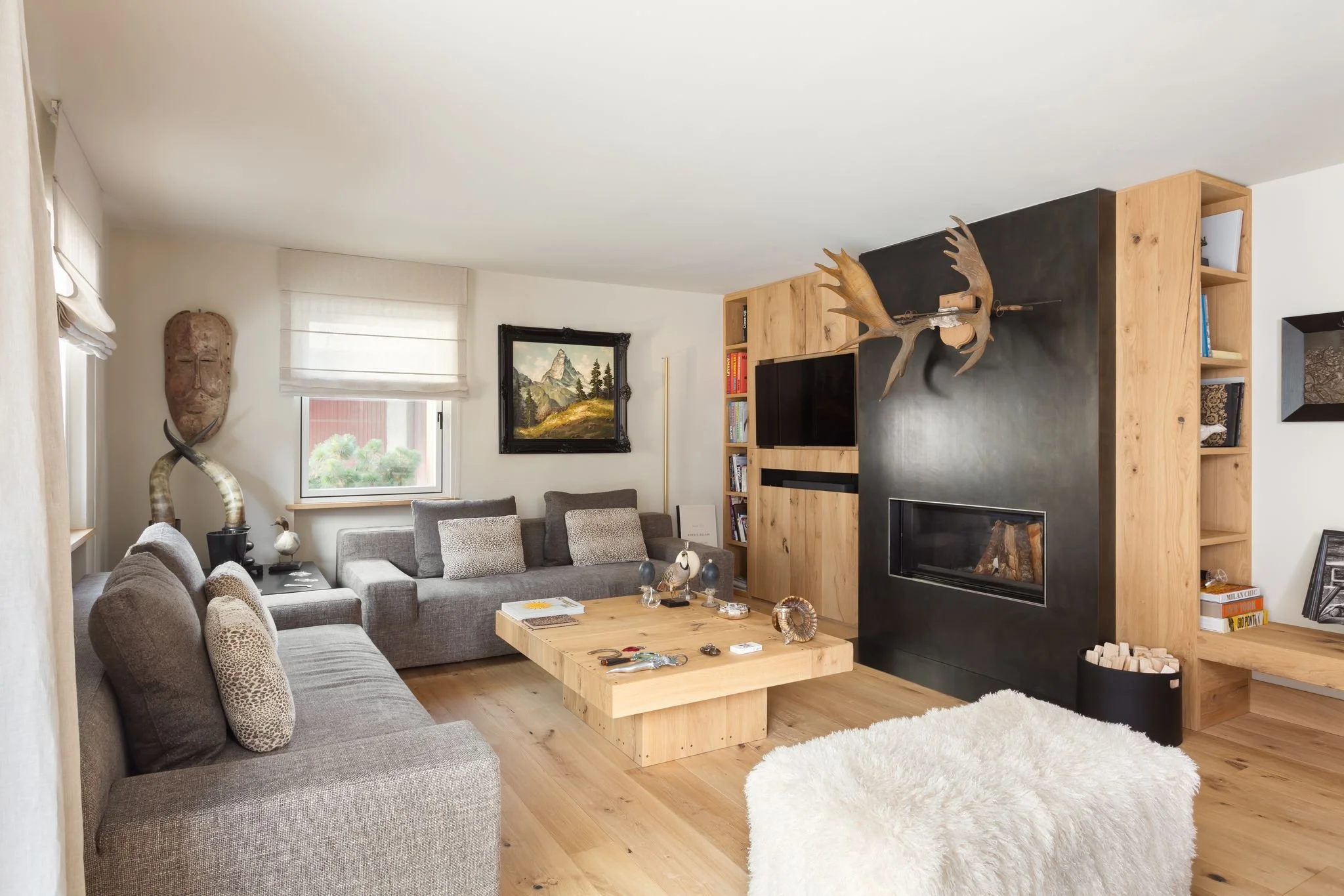 Soggiorno arredato in stile moderno con divani grigio, tavolino in legno naturale, camino a parete in bianco e nero, decorazioni di corno e statuetta, scaffali con libri e decorazioni, quadro di paesaggio sulle pareti, tappeto bianco e finestre con tende chiare.