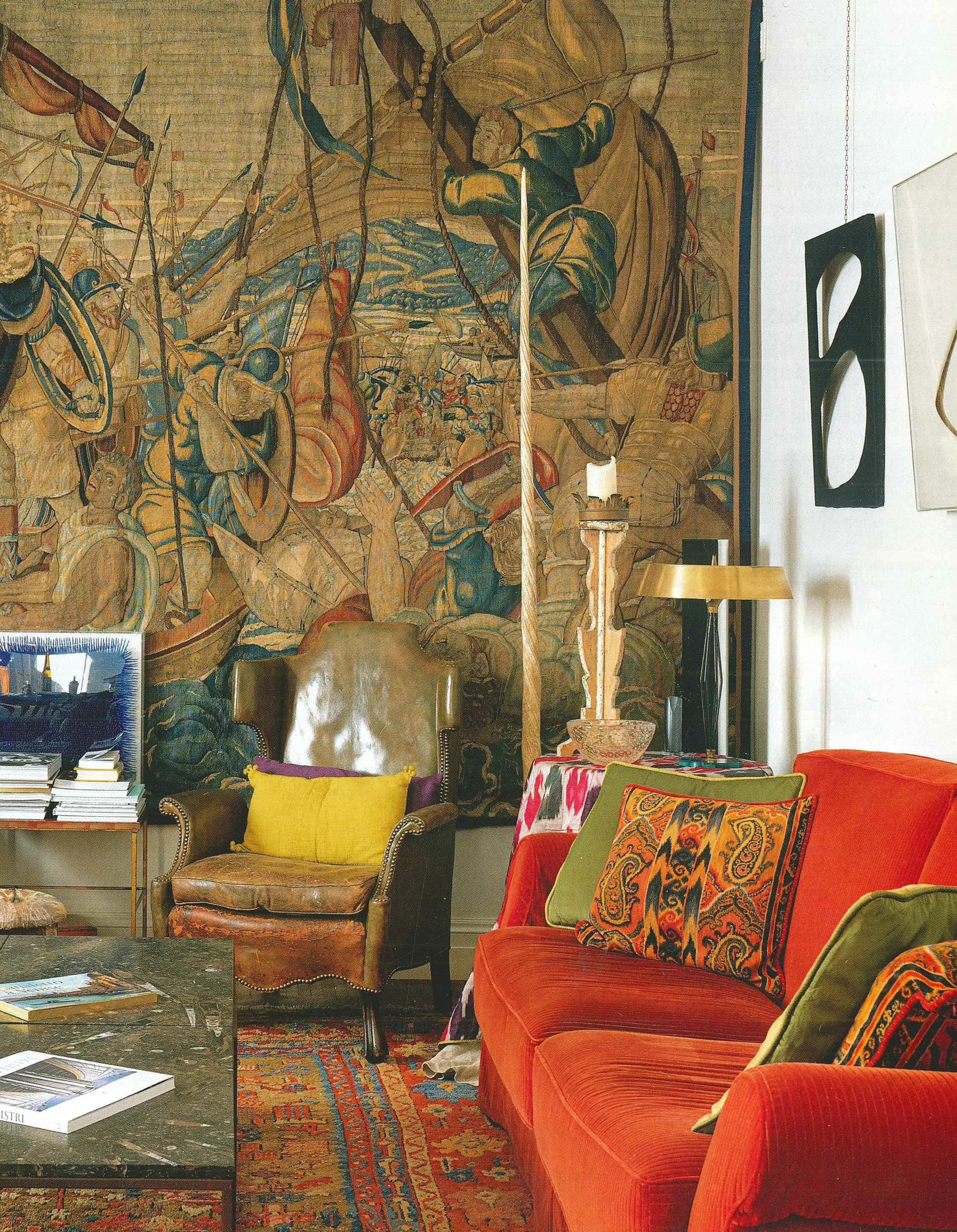 Soggiorno con divano arancione, poltrona in pelle marrone, lampada e tappeto con motivi geometrici e floreali, grande arazzo con scene storiche sul muro, scatola con libri e riviste a sinistra.