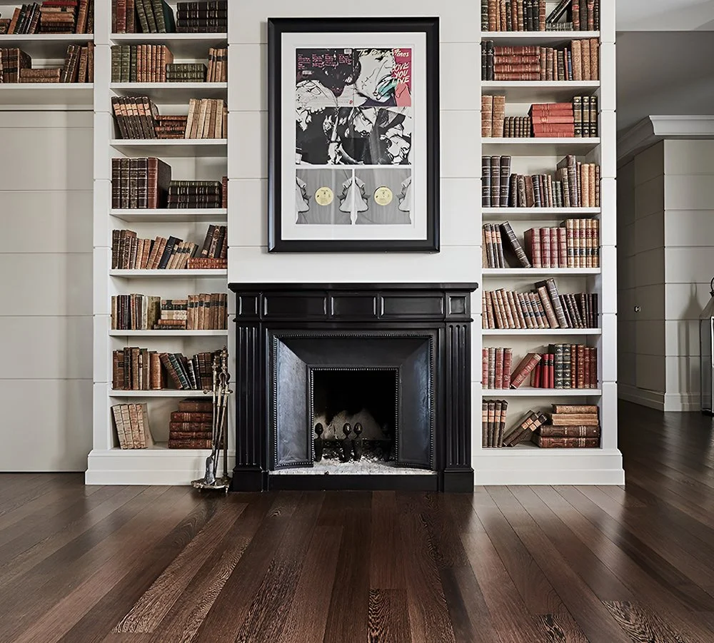 Stanza con pareti rivestite di scaffali pieni di libri antichi, con un camino nero e sopra un quadro con arte moderna; pavimento in legno scuro.