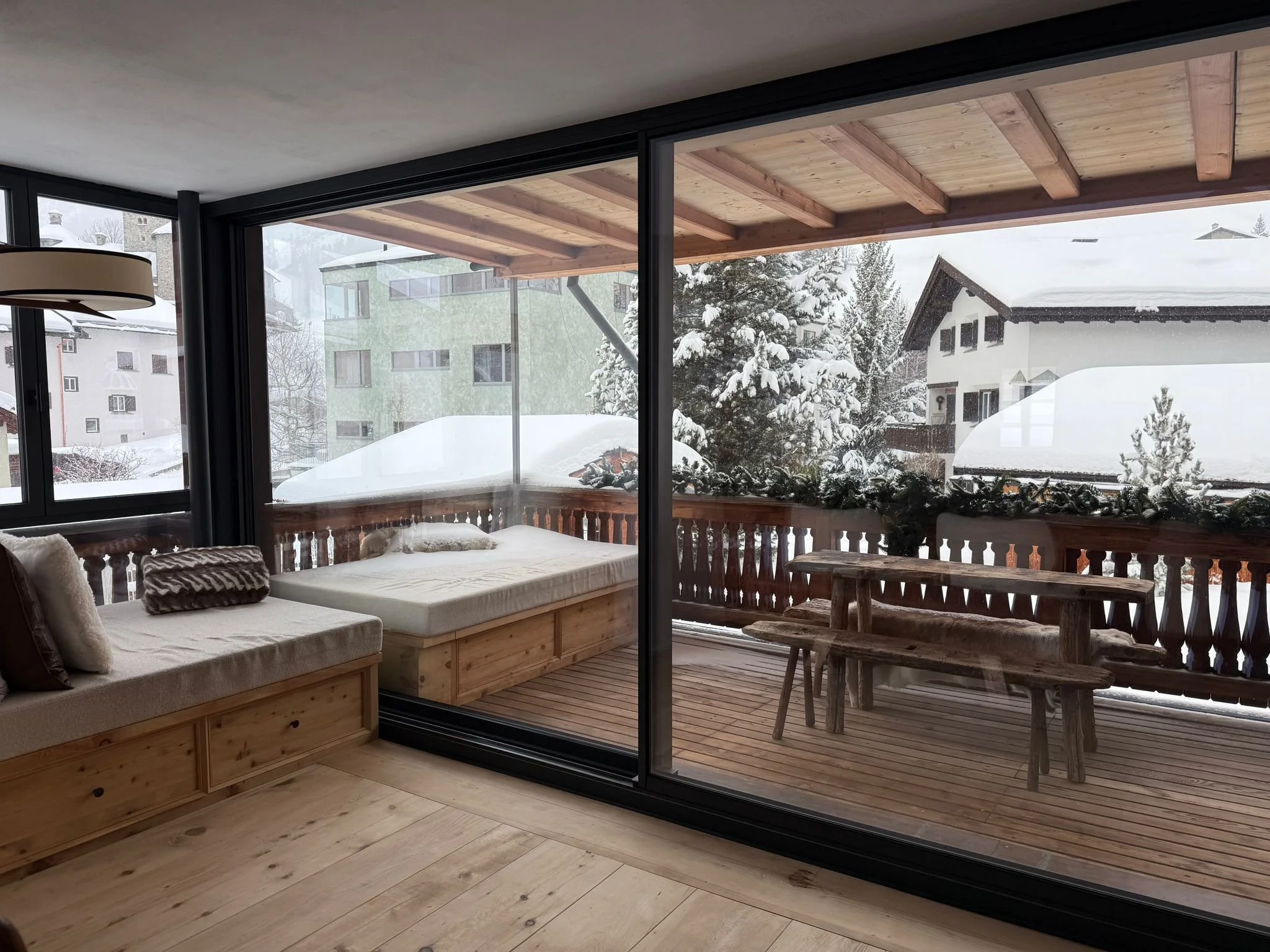 Interno di una stanza con grandi finestroni che si affacciano a un balcone coperto di neve. Sul balcone, tavolo e panca in legno, con alberi e case innevate all'esterno.