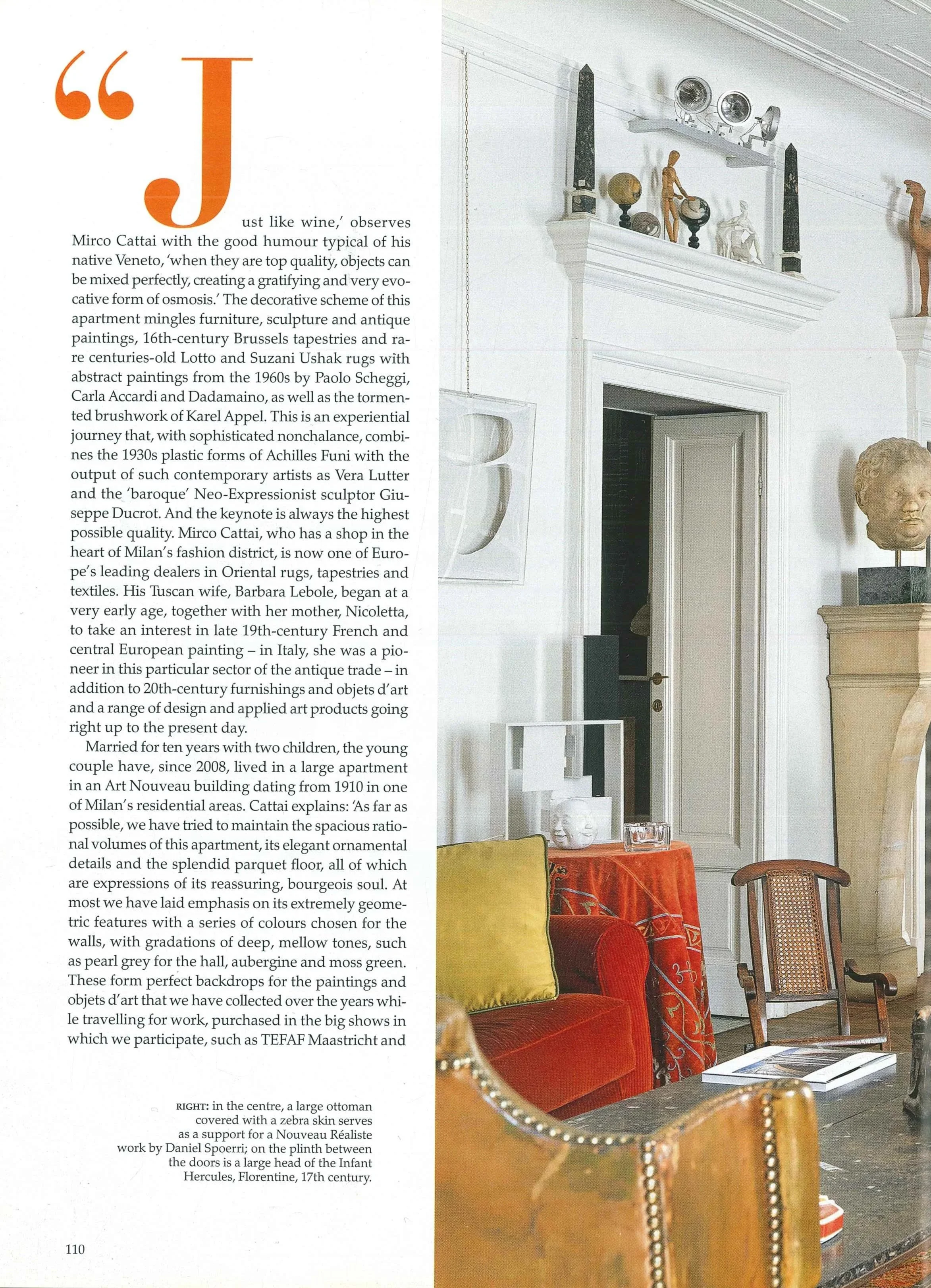 Interno di un appartamento con decorazioni d'antiquariato, sculture, teleri e mobili vintage, tra cui un divano rosso, una poltrona in paglia, e oggetti d'arte su mensole e tavola.