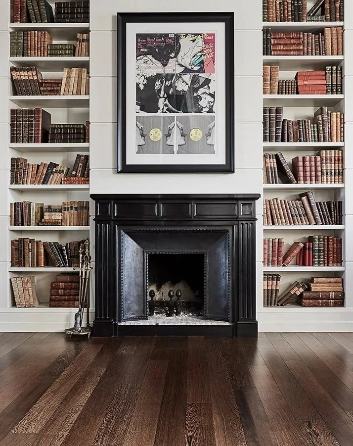 Stanza con librerie piene di libri alle pareti ai lati di un camino nero, con un quadro sopra e un pavimento di legno.