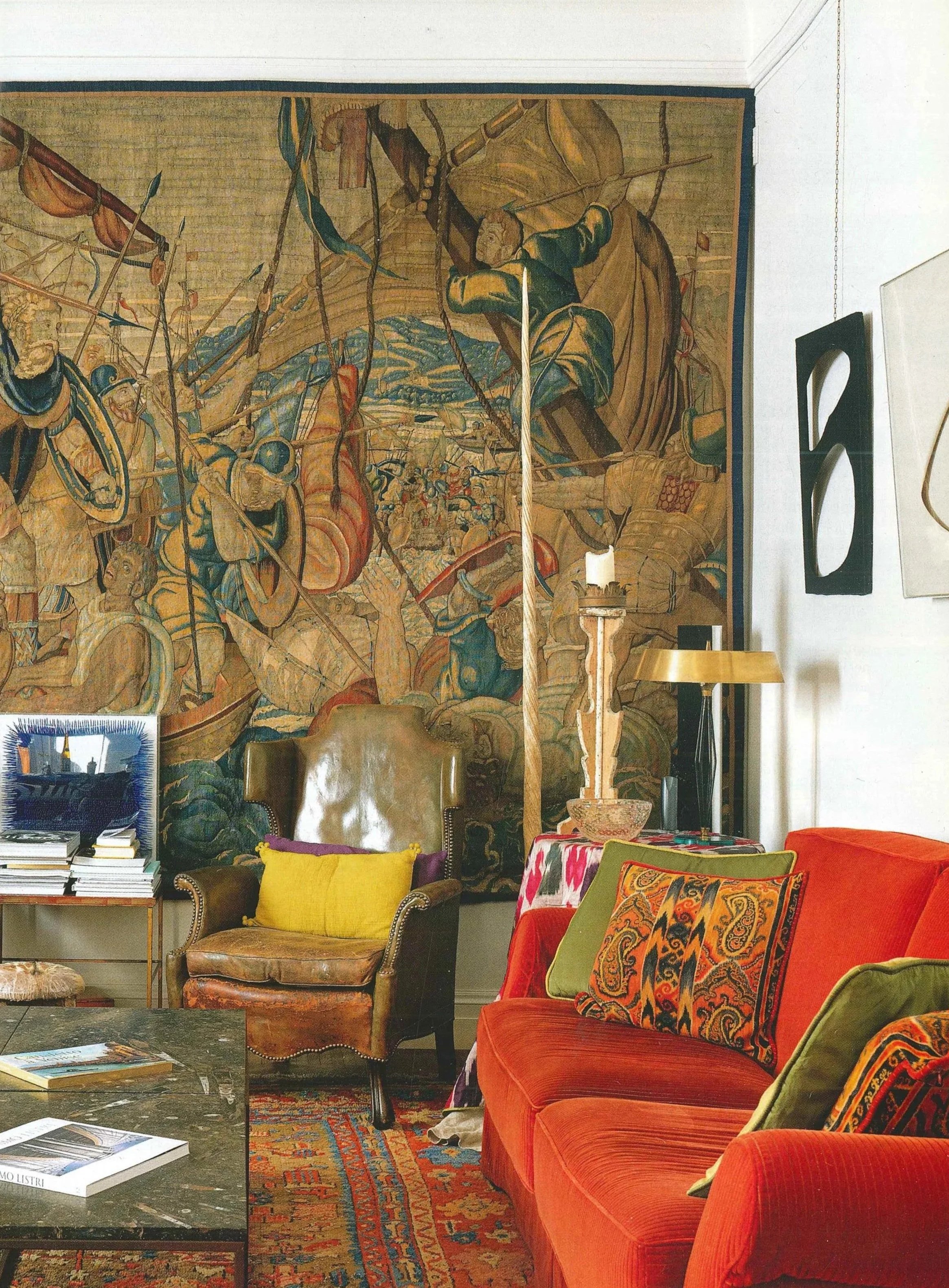 Interno di un soggiorno con un grande arazzo alle spalle, un divano rosso con cuscini vari, una poltrona marrone, un tavolino con riviste, una lampada da terra e una lampada da tavolo, decorazioni artistiche sulla parete.