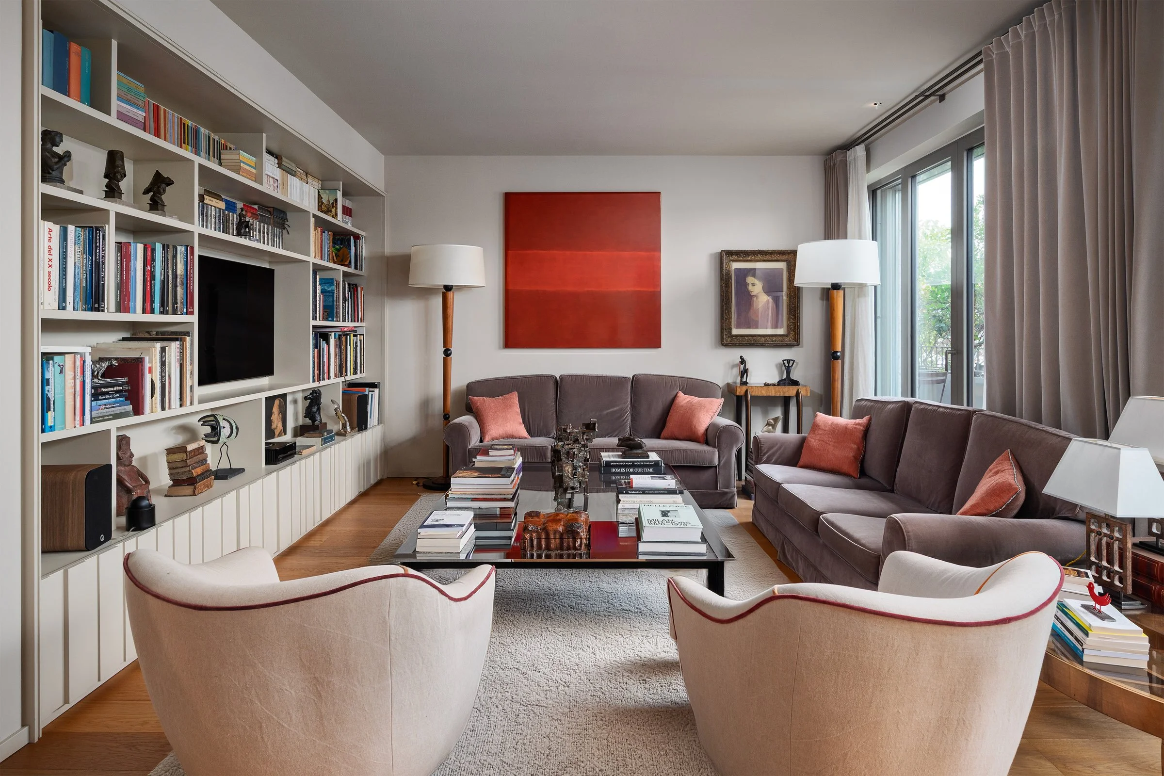 Soggiorno con divani grigi, poltrone beige, libreria bianca, tappeto beige, tavolino con libri e oggetti decorativi, lampade da terra, quadri e tende color crema.
