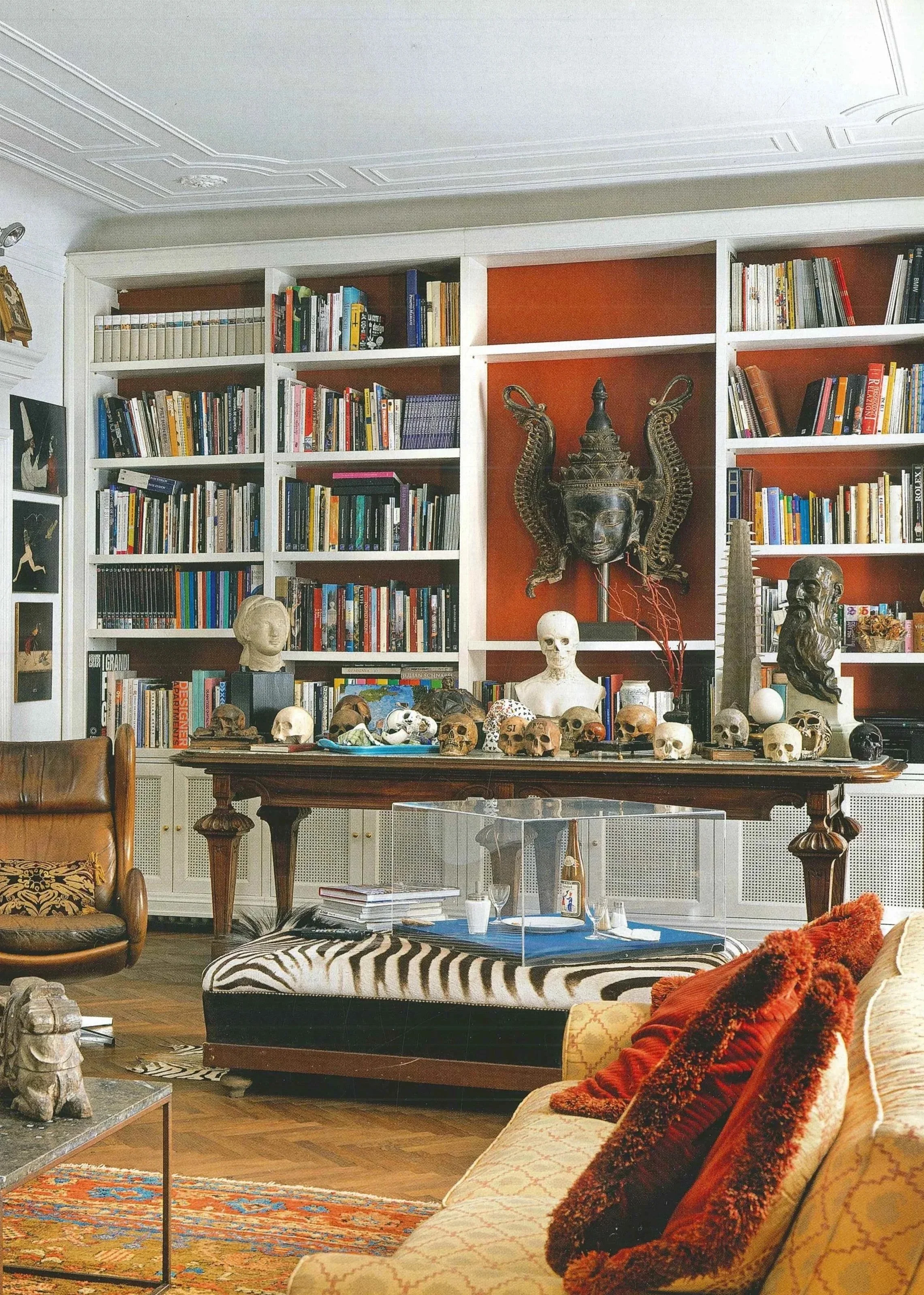 Un soggiorno con libreria a parete piena di libri, busti e arte decorativa, con divani e tavolino decorato con oggetti, skulls, e un animale imbalsamato.