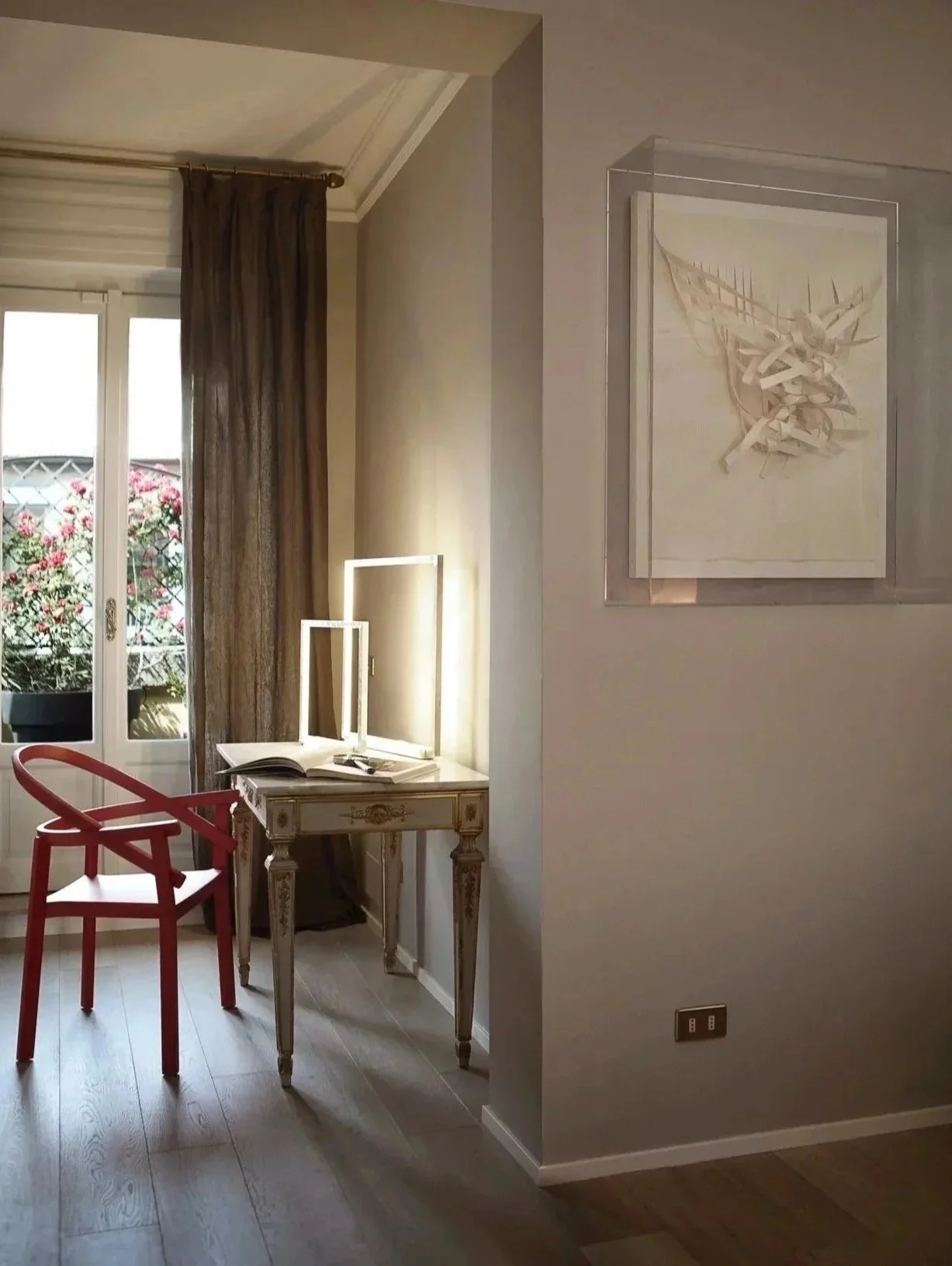 Interior di una stanza con pareti color pesca chiaro, un tavolo antico con cassetti e decorazioni dorate, una sedia rossa e una finestra con tende di velluto marrone che lascia entrare la luce naturale, un quadro astratto con tonalità neutre e un balcone con piante e fiori visibili dall'esterno.