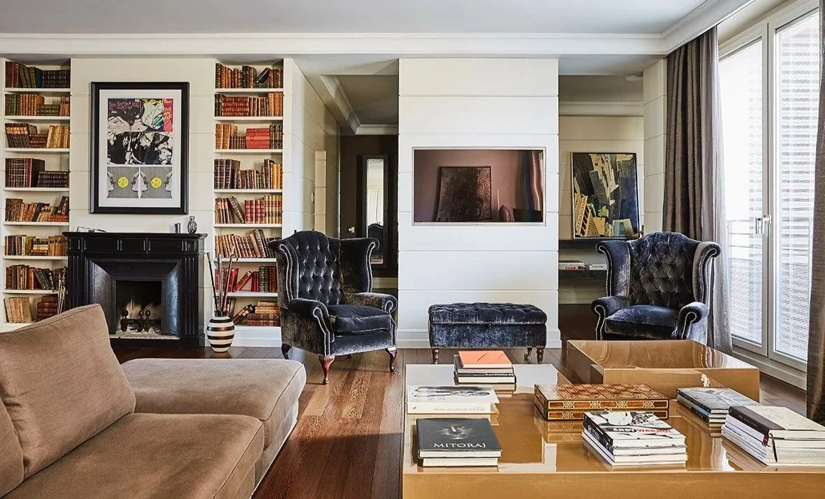 Soggiorno elegante con pareti rivestite di scaffali pieni di libri, due poltrone nere con schienale alto, un divano beige, un camino nero, una parete con un'installazione artistica, grandi finestre con tende, e un tavolino con libri e decorazioni.