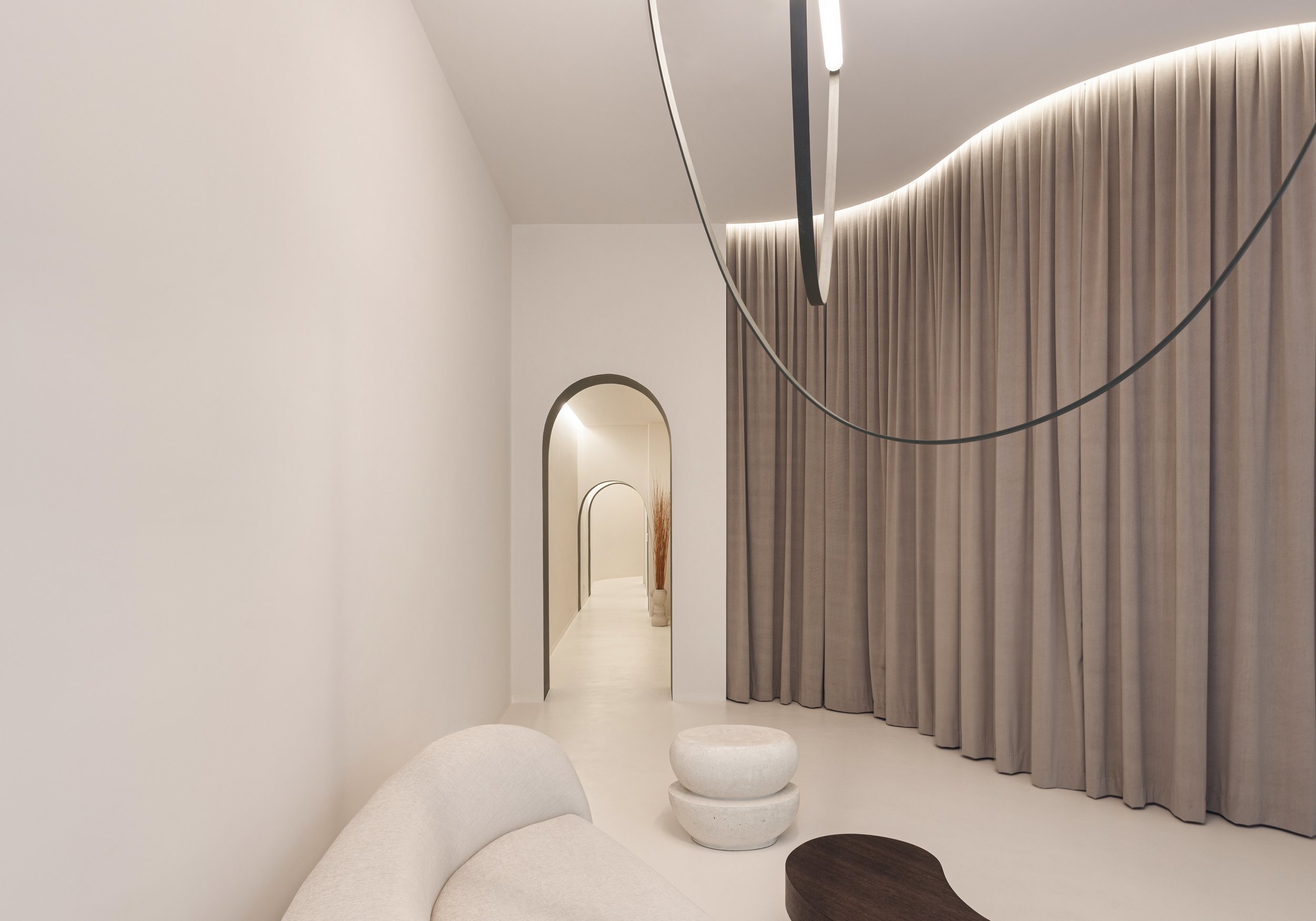 Interno minimalista con pareti bianche, grande tende beige, soffitto curvo con luci indirette, divano bianco, tavolino di pietra, decorazioni semplici e arco decorativo.