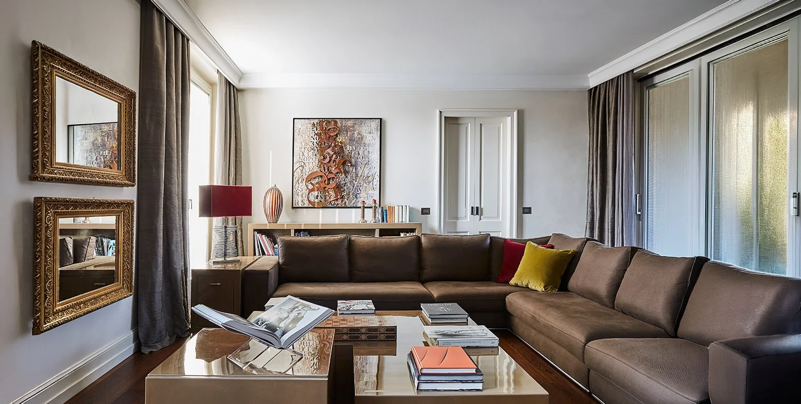 Soggiorno con divano marrone, tavolino con libri, pareti decorate con quadri e specchi, finestre con tende e luce naturale.