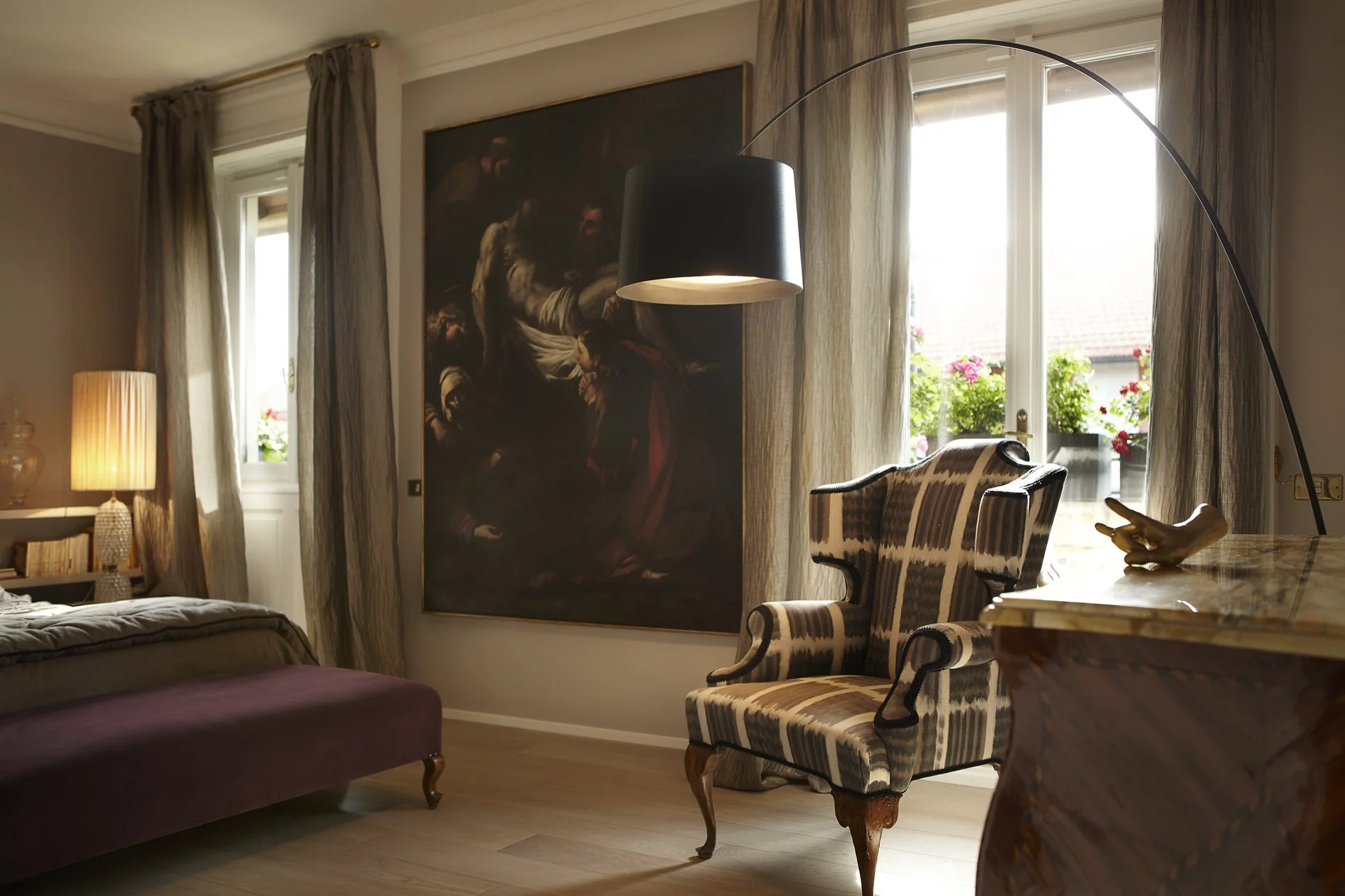 Interno di una stanza con un divano, una sedia a braccioli con tessuto a righe, un quadro grande e una finestra con vasi di gerani.