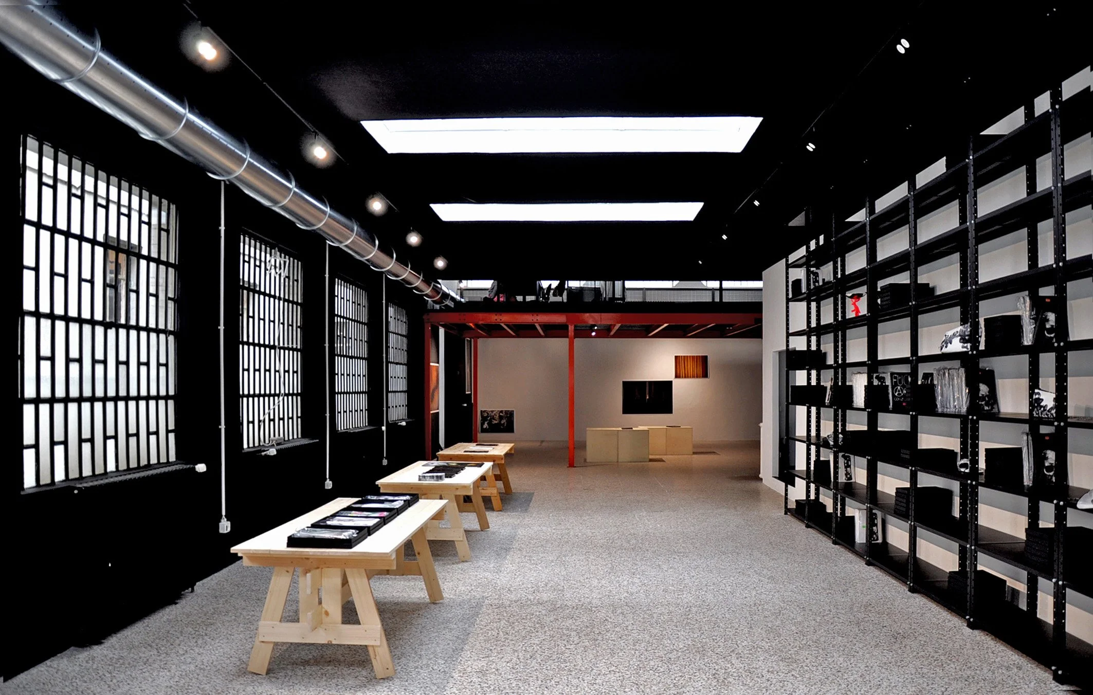 Interno di una galleria d'arte moderna con pareti nere, scaffali neri pieni di oggetti e tavoli di legno con cataloghi o libri, illuminato da luci sopra e lucernari sul soffitto.