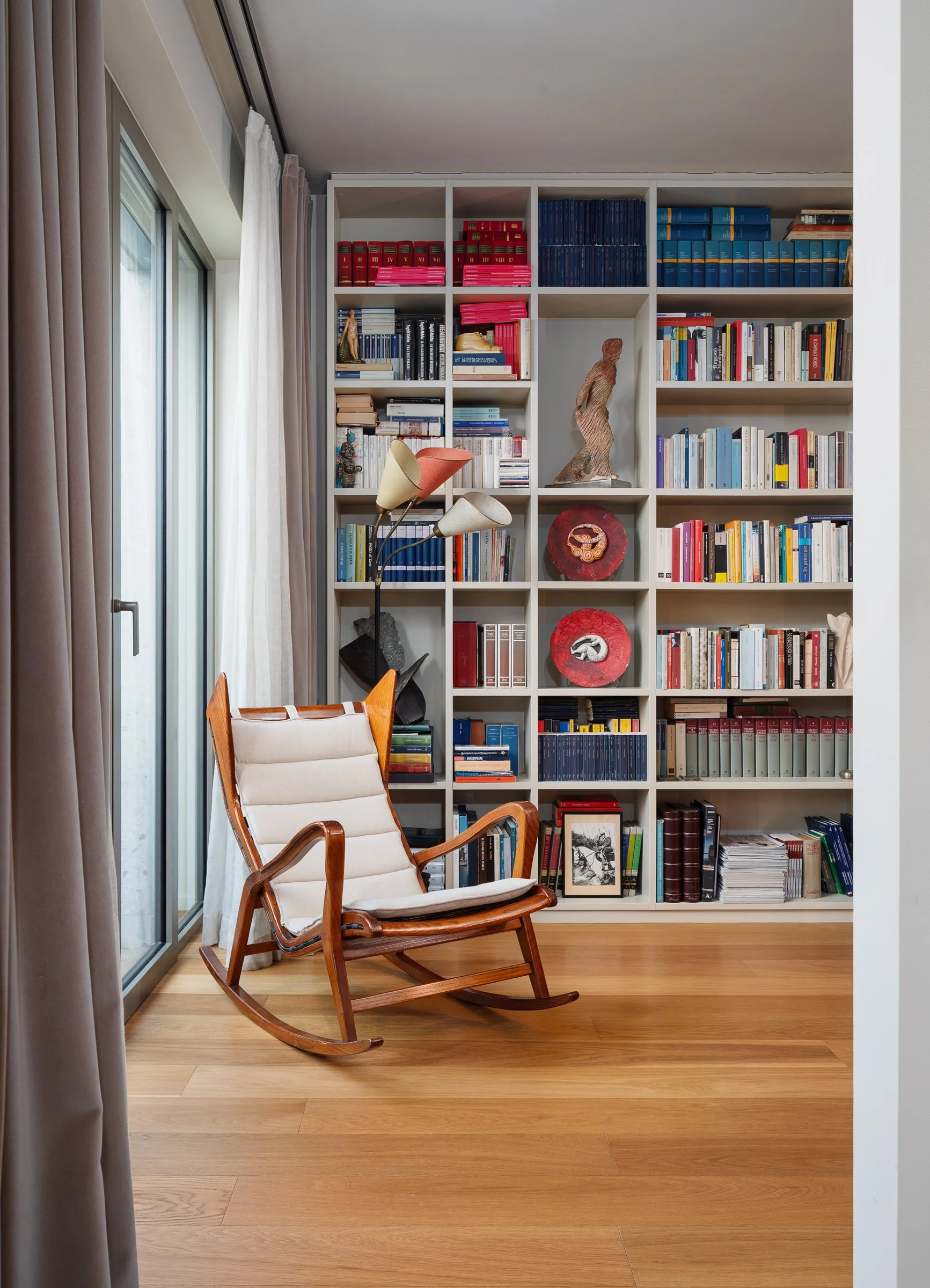 Soggiorno con poltrona a dondolo di legno chiaro, libreria bianca piena di libri e decorazioni, tappeto, e grandi finestre con tende beige e persiane.