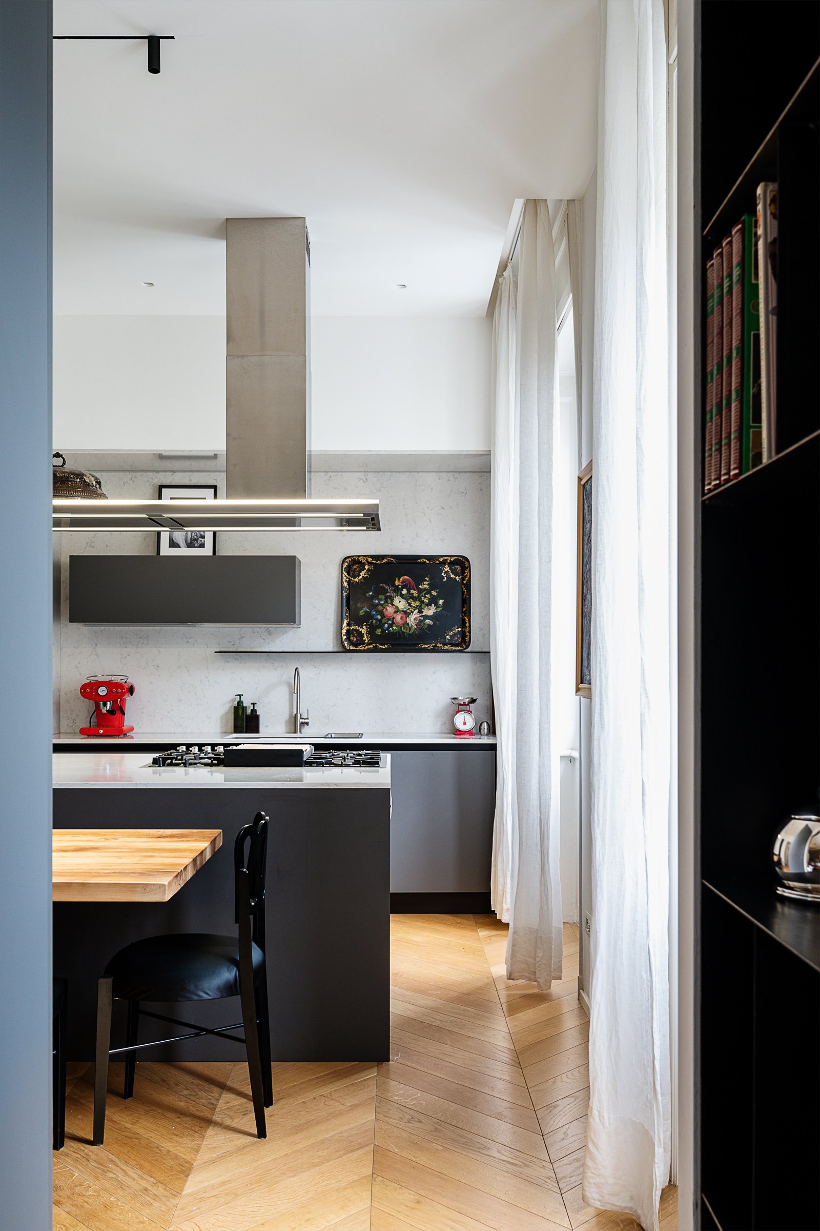 Cucina moderna con pareti bianche, armadietti grigi, lavabo, piano cottura e una moka in acciaio. C'è anche un quadro con decorazioni floreali sulla parete e tende bianche alle finestre.