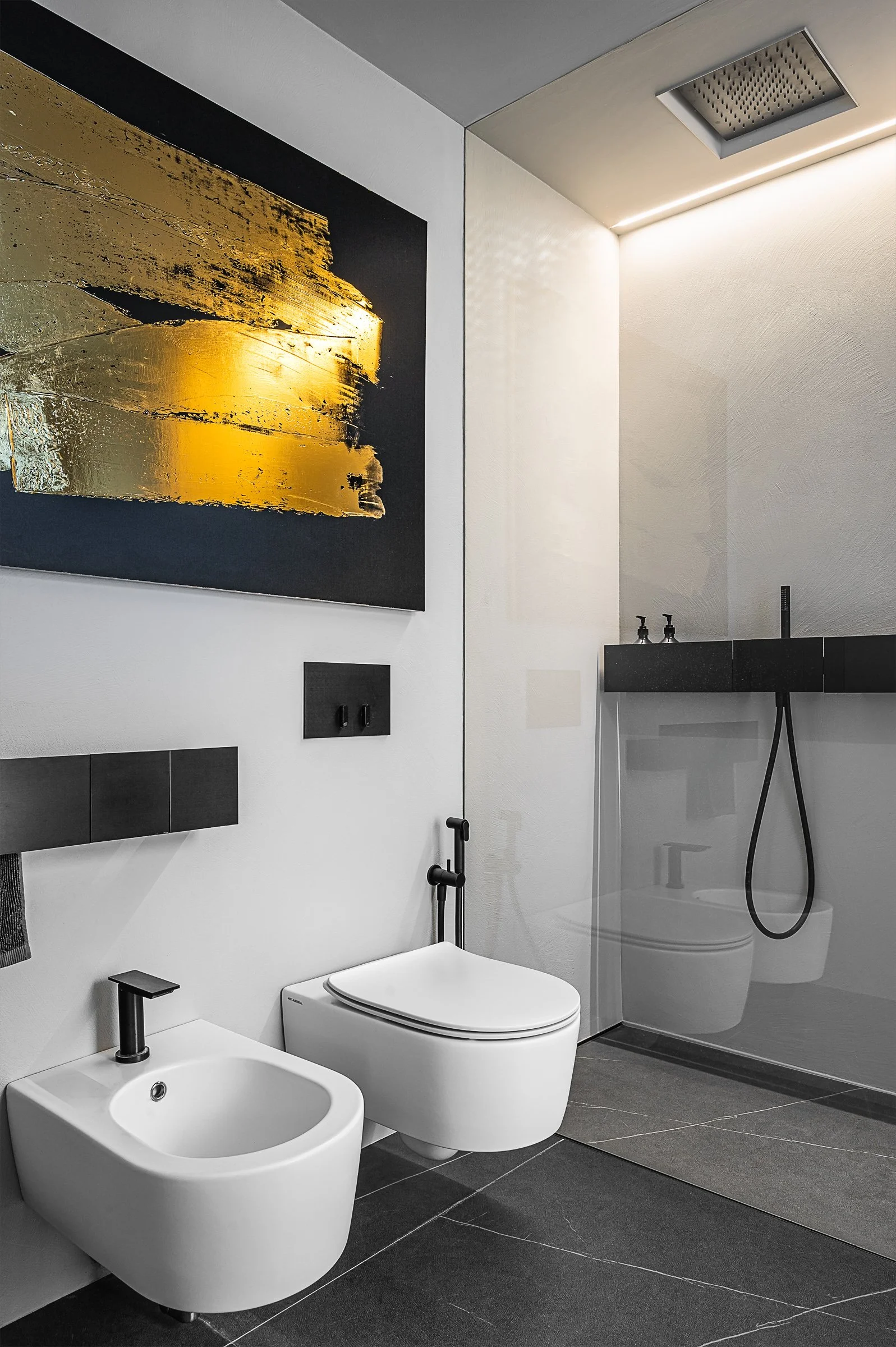 Bagno moderno con tazza wc e bidet bianchi, pareti bianche, pavimento in piastrelle nere, vasca con soffione doccia e parete divisoria in vetro, quadro astratto con toni dorati e neri.
