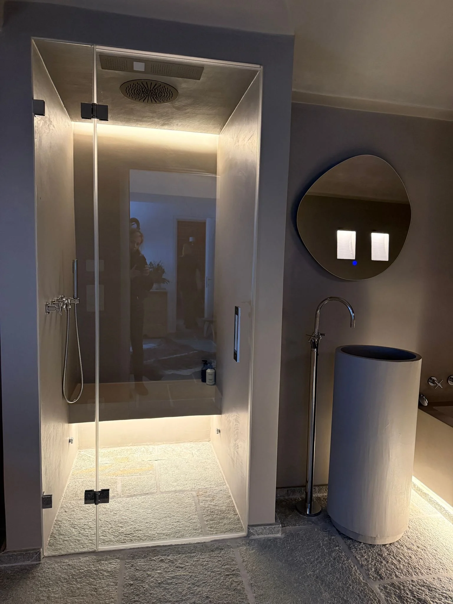 Doccia moderna con pareti di vetro trasparenti, soffitto con soffione grande, pareti in legno chiaro, pavimento in pietra, vicino a uno specchio rotondo e un lavabo cilindrico in ceramica, con un rubinetto a fianco.