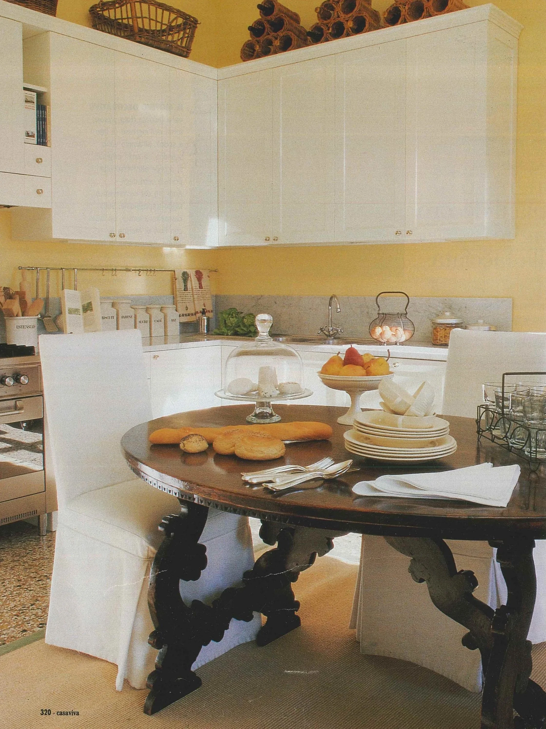 Interno di una cucina con tavolo rotondo, piatti, utensili e decorazioni, pareti gialle, armadi bianchi e ripiani con oggetti, e una cucina a gas