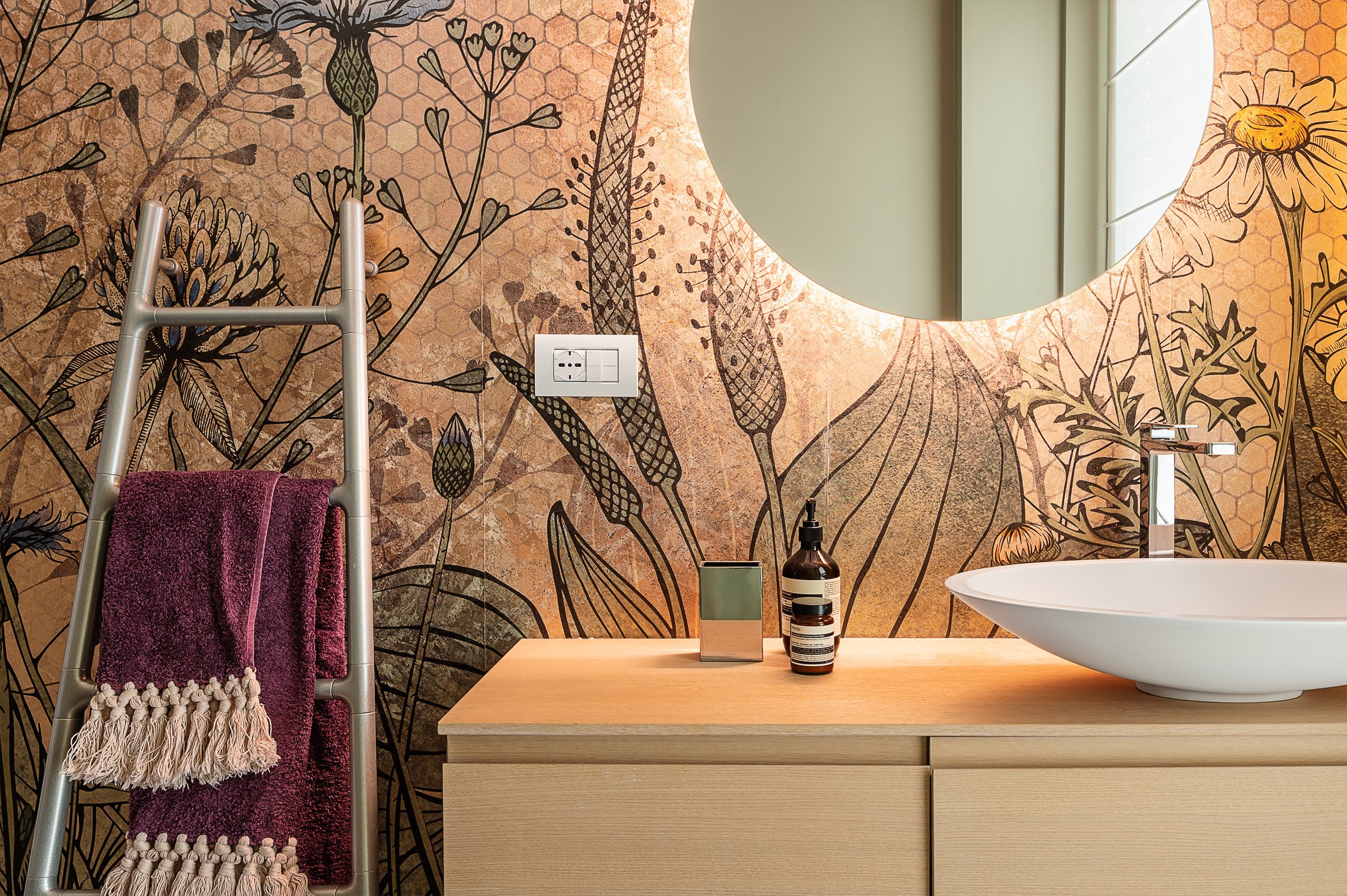 Bagno con parete decorata con motivo floreale e sfondo a motivi esagonali, specchio rotondo sopra un lavabo bianco a ciotola, finestra che lascia entrare la luce naturale, una scala con asciugamani bordeaux appesi, e oggetti come un dispenser di sapone, un portasciugamani e un contenitore in metallo sulla superficie in legno del mobile.