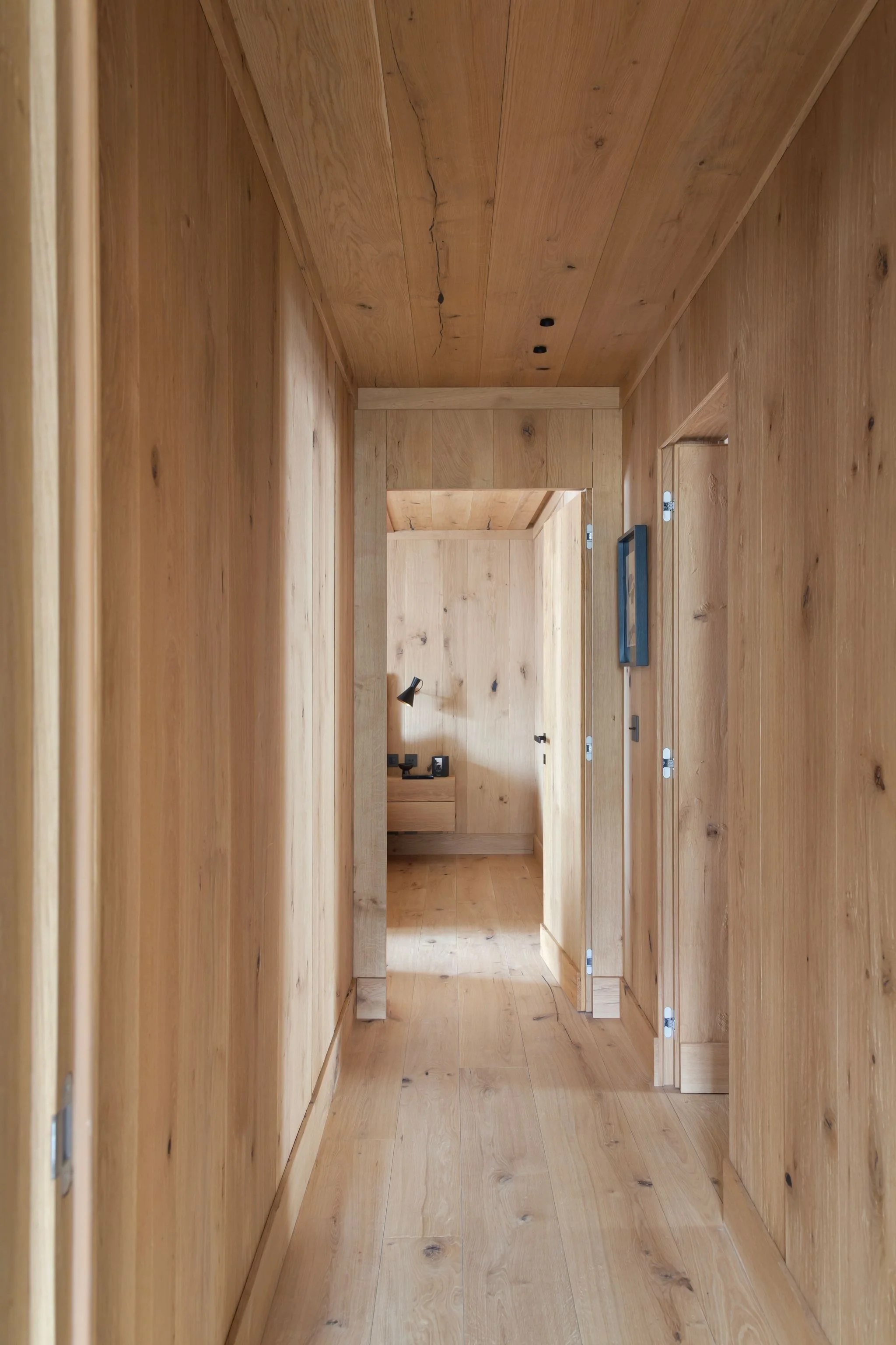 Corridor di casa in legno con pareti e pavimenti in legno chiaro, vista fino a una stanza con mobile e lampada da scrivania.