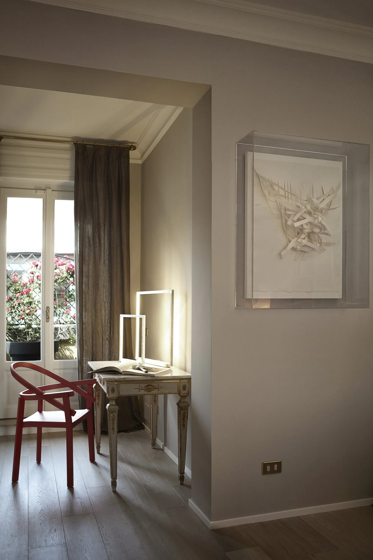 Angolo di una stanza con un tavolo decorato, una sedia rossa e una finestra con tende marroni, con balcone e piante floreali all'esterno.