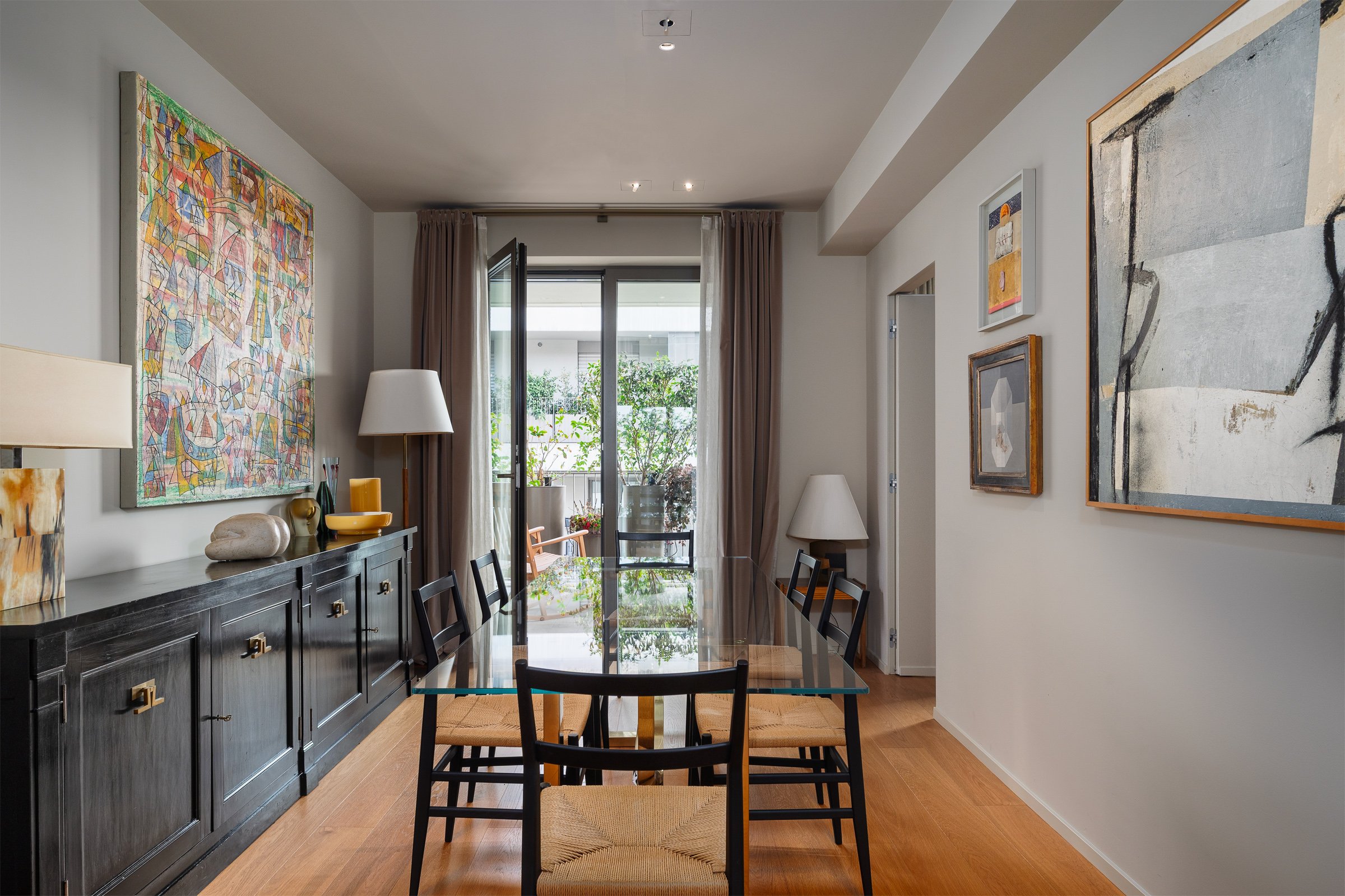 Interno di una sala da pranzo con tavolo in vetro, sedie nere con sedili di paglia, grande armadio nero con decorazioni in ottone, e pareti decorate con dipinti moderni.