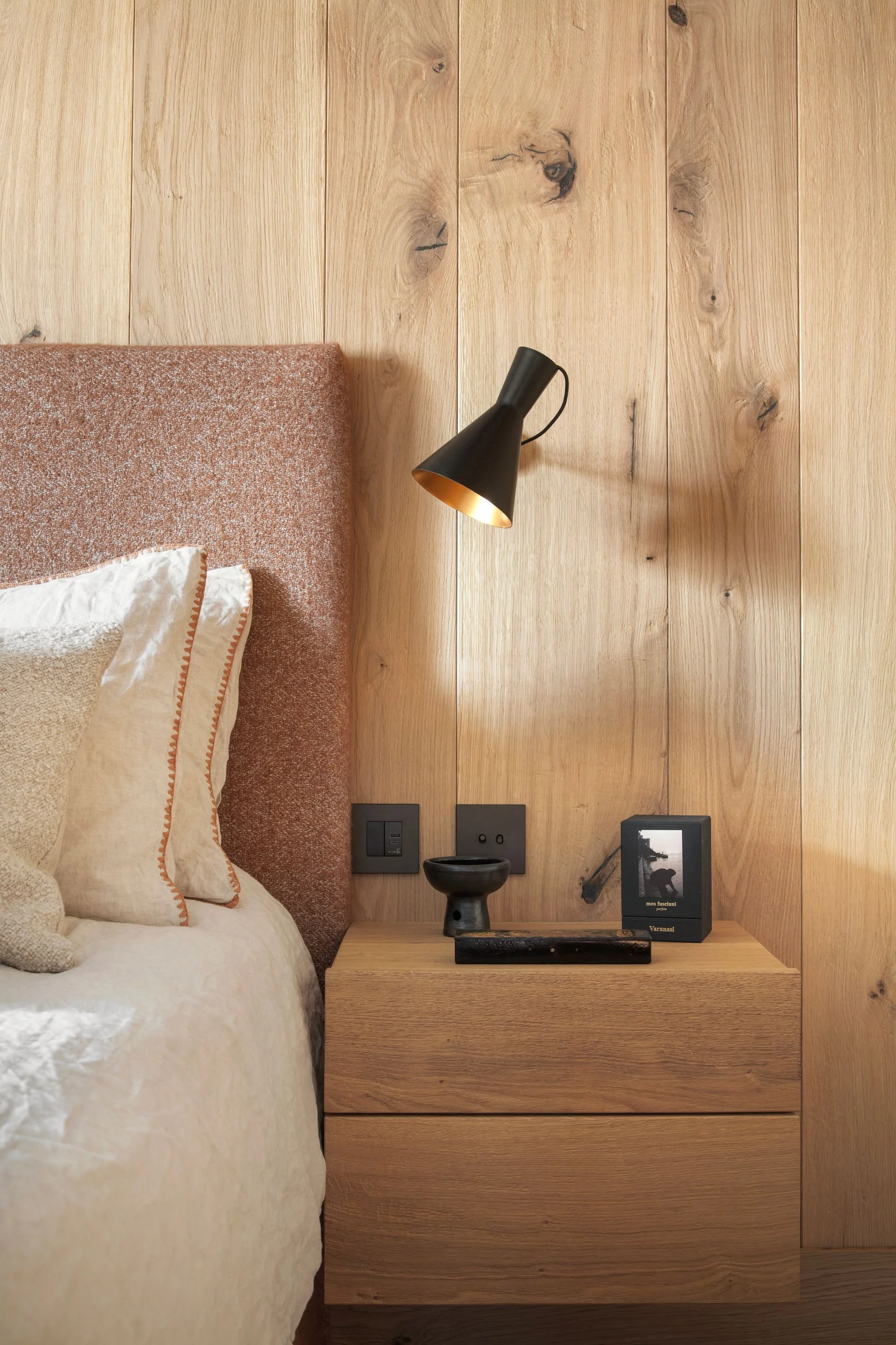 Sulla comodino in legno ci sono un quadro con foto e una ciotola nera, sul muro di legno una lampada moderna nera e due prese di corrente