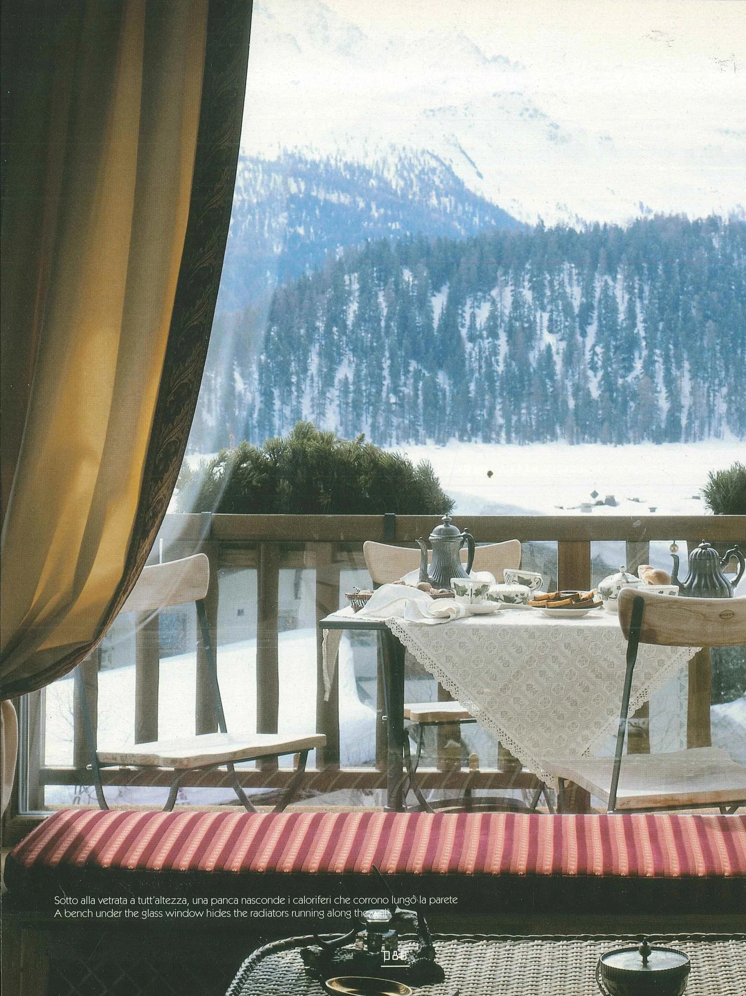 Vista di una tavola imbandita sul balcone con una scena montana innevata sullo sfondo, e il soggiorno che guarda fuori. C'è una sedia, un tavolino con tazze, teiere e piatti, e una finestra con tende dorate.