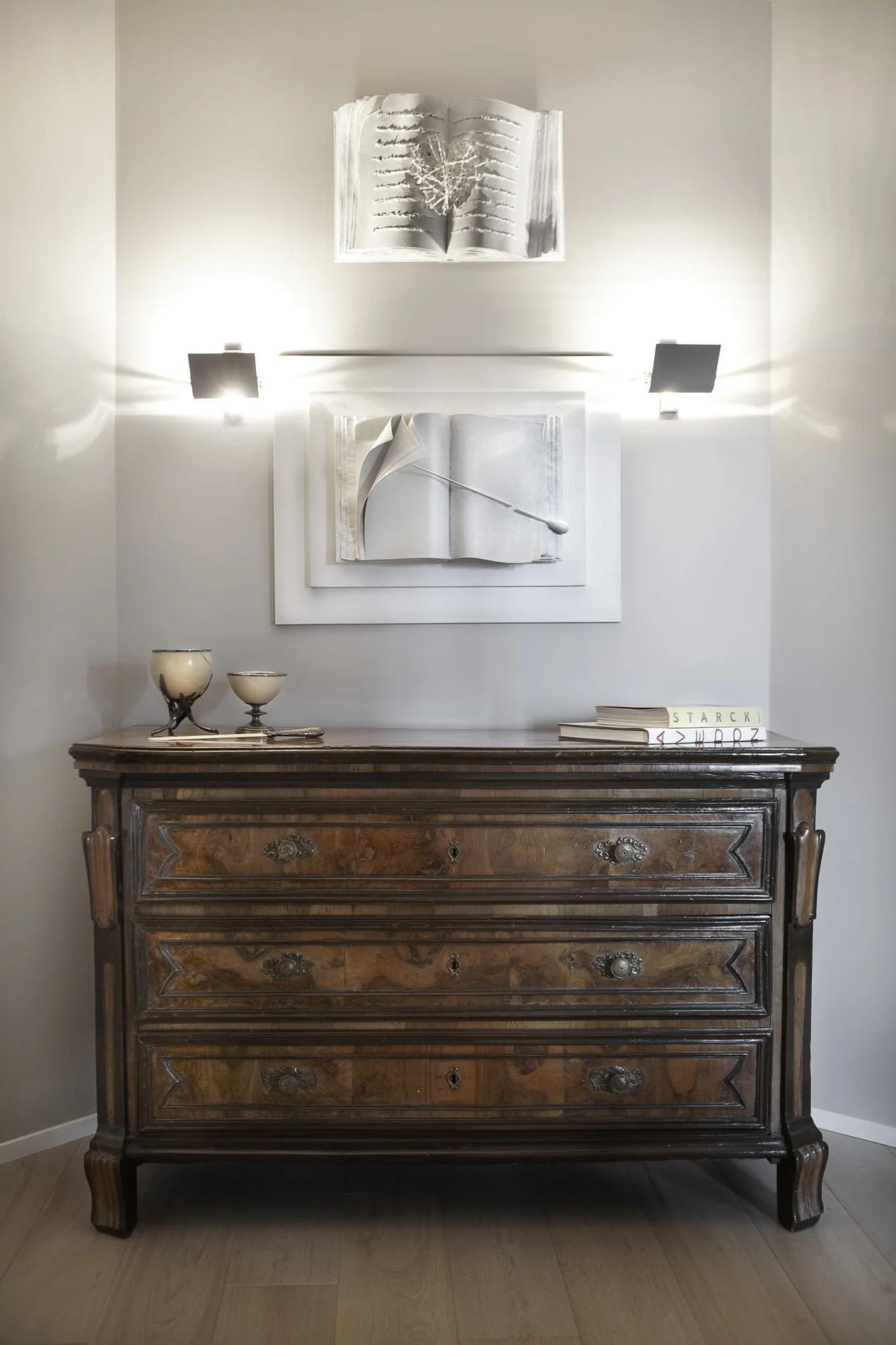 Una credenza di legno antico con tre cassetti, sopra alcune ciotole e libri, con decorazioni in bianco alle pareti e due lampade luminose.
