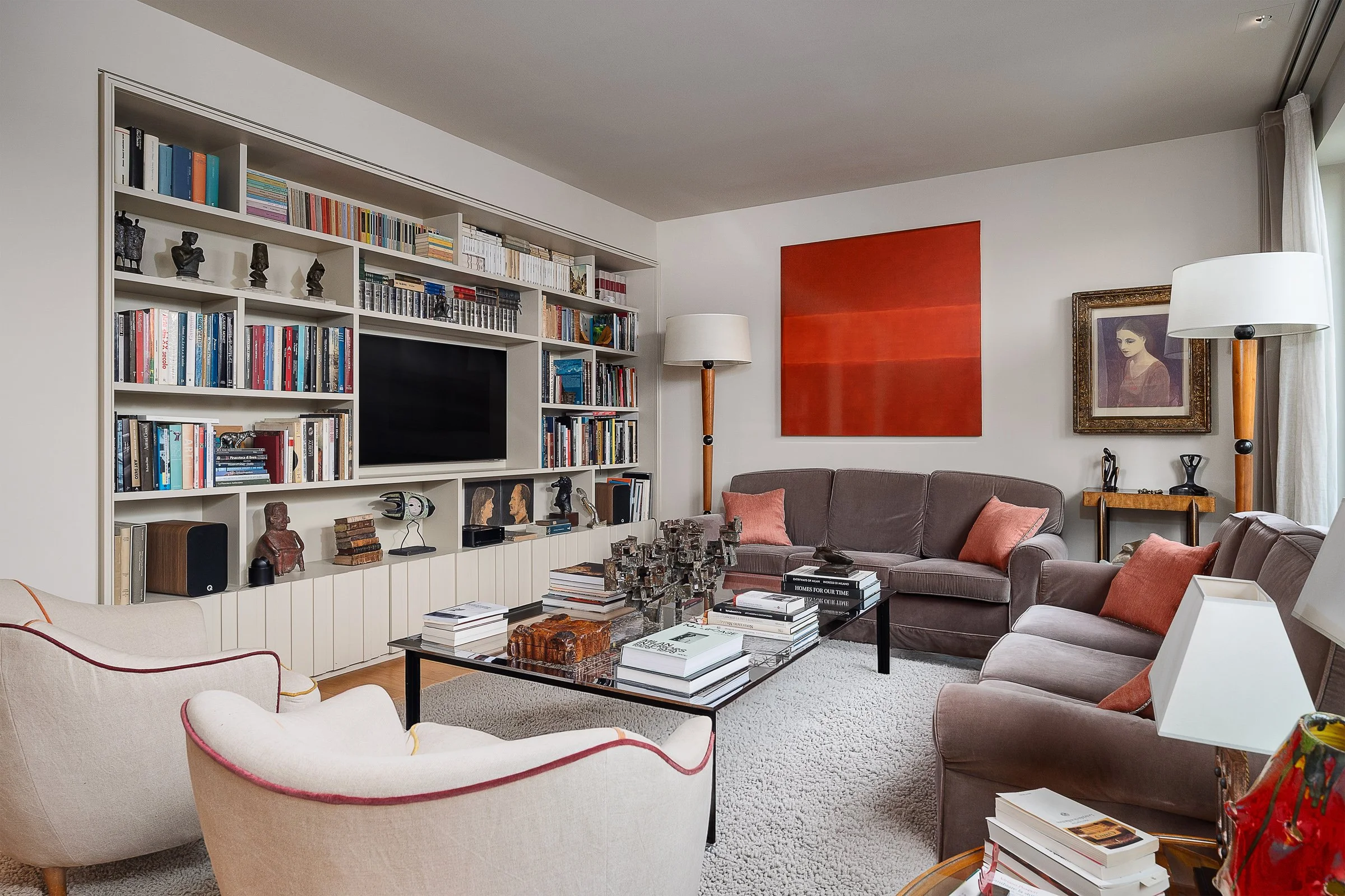 Soggiorno elegante con libreria bianca a parete, divano marrone con cuscini rosa, sedie bianche, tavolino con libri e decorazioni in metallo, quadri e lampade