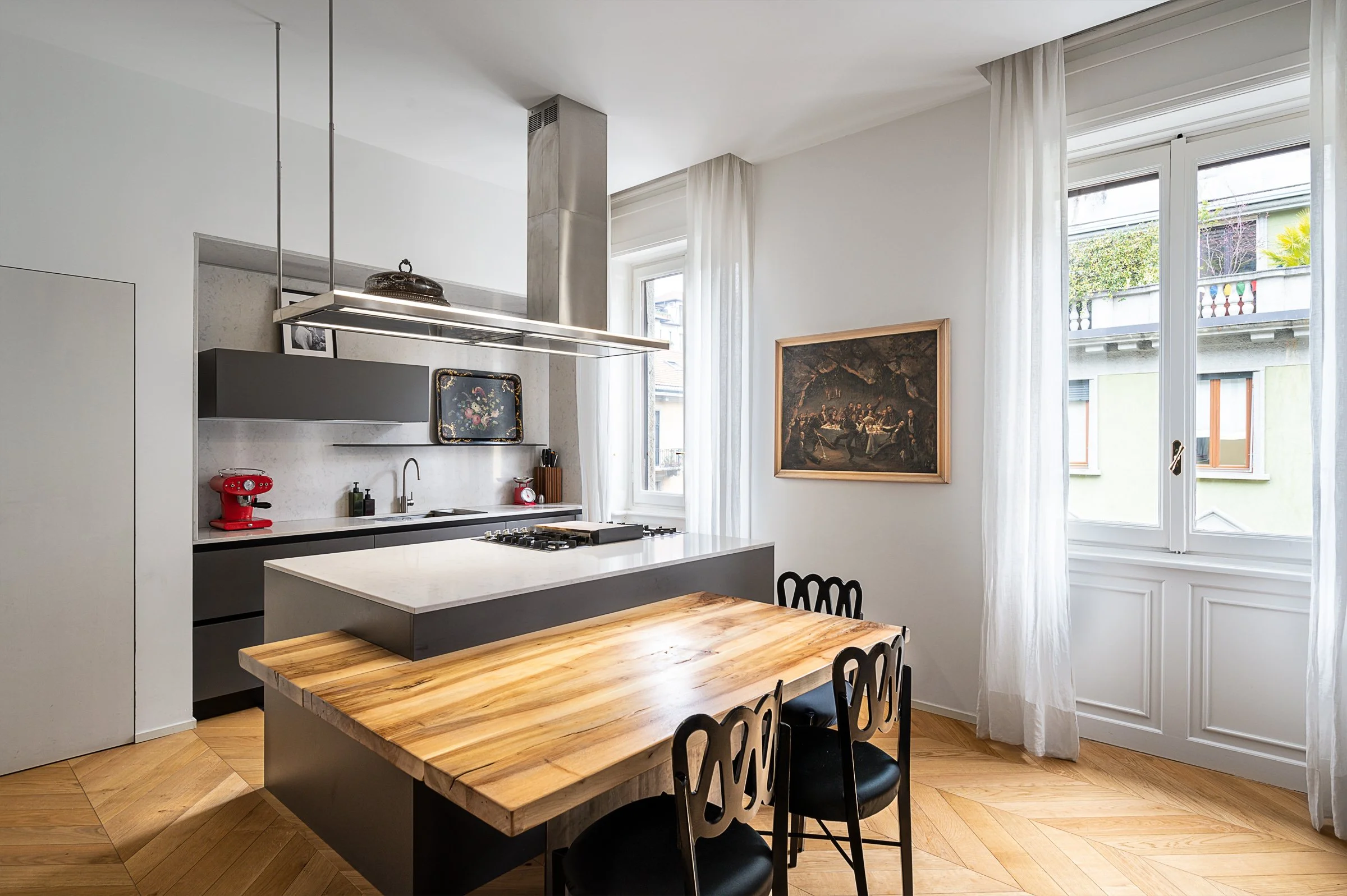 Cucina moderna con tavolo in legno, sedie nere, pareti bianche, grande finestra con tende bianche, dipinto appeso alla parete.