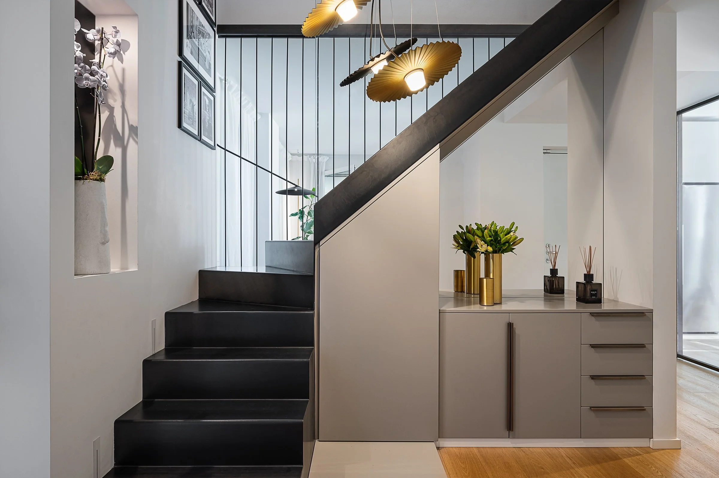 Interno moderno di casa con scala nera, decorazioni floreali in vasi dorati e oggetti di design, pareti bianche.