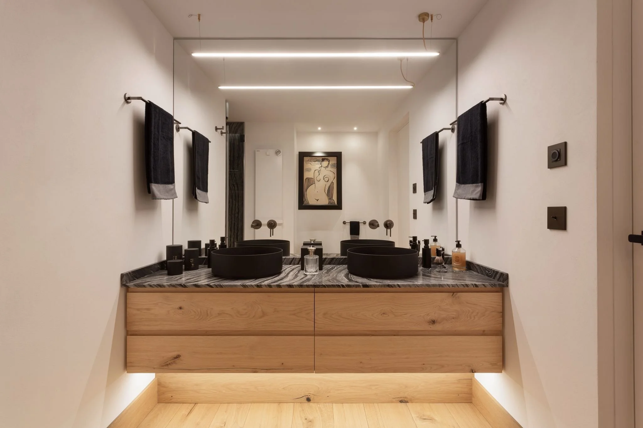 Bagno moderno con due lavabi neri su un piano di marmo, specchio grande sopra i lavabi, asciugamani neri appesi alle pareti, quadri di arte moderna alle pareti, pavimento di legno chiaro.