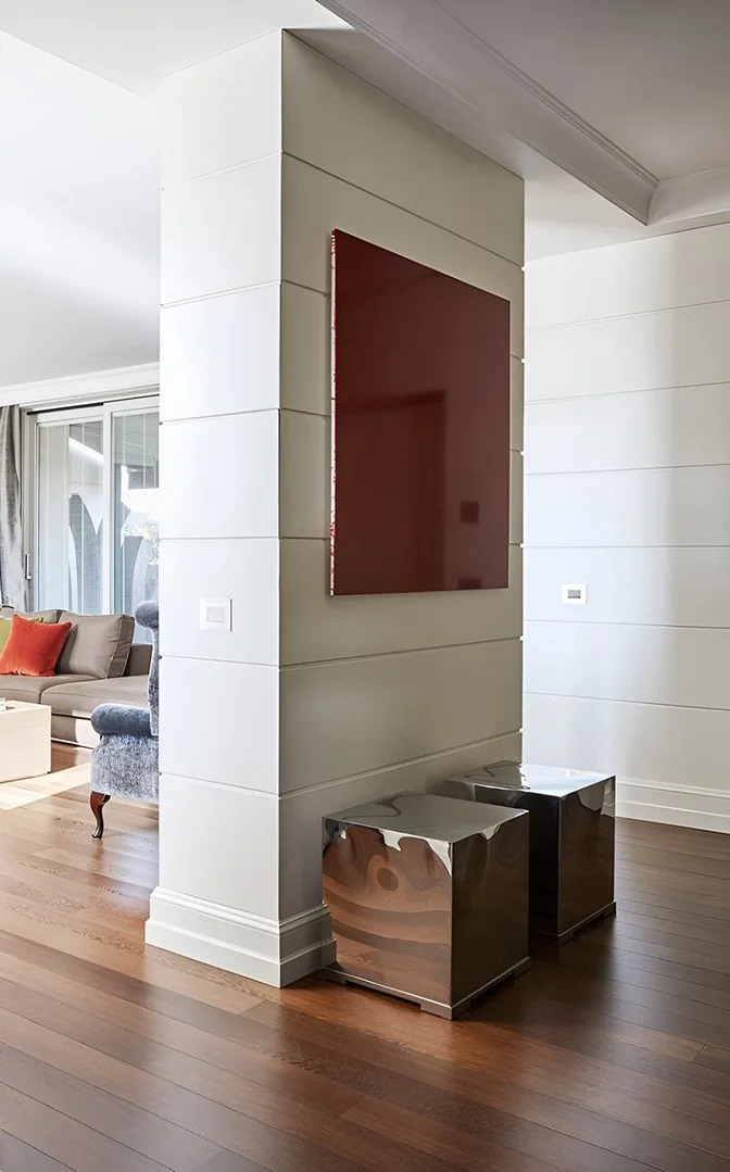 Interno di una casa moderna con pareti bianche, un quadro rosso a parete, due tavolini di metallo lucido e un'area soggiorno con divani e cuscini.