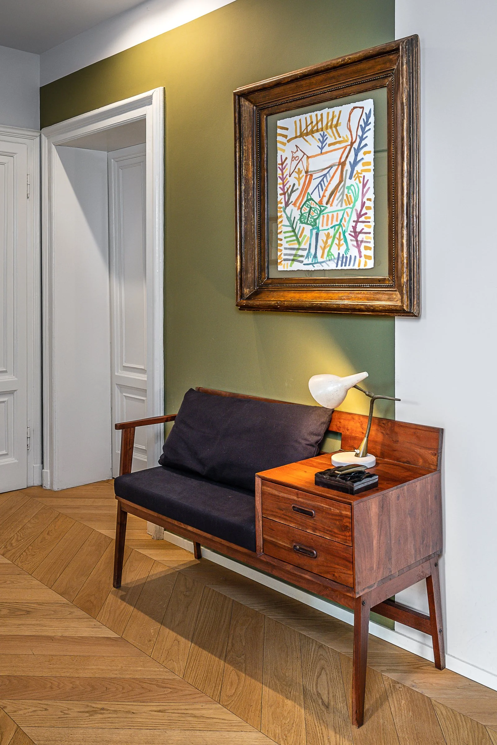 Angolo di un interior con una panchina di legno con schienale nero, una lampada da tavolo bianca in parte appoggiata sul bracciolo, un mobiletto con due cassetti, un quadro con disegno stilizzato di cervi e natura in cornice in legno.