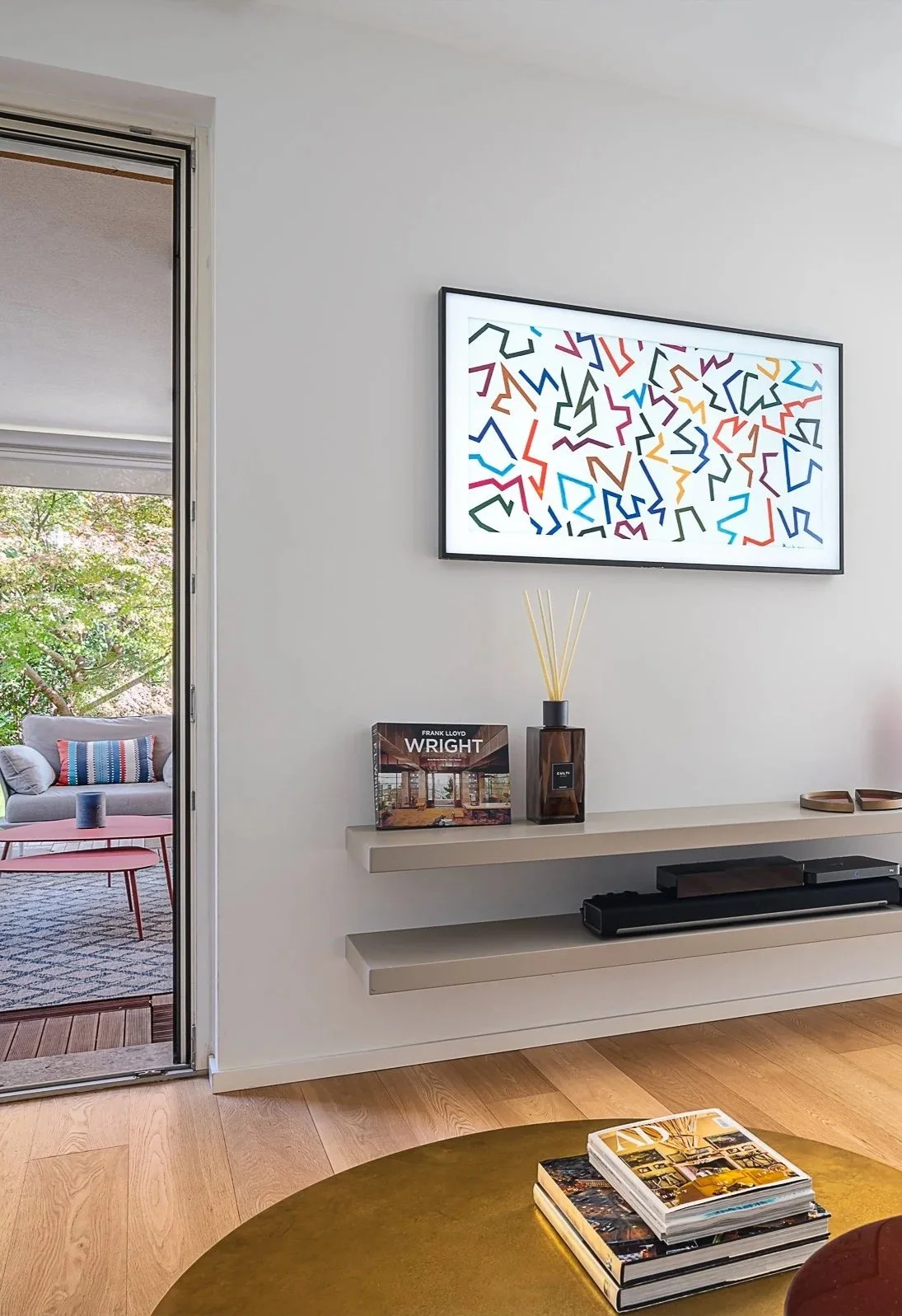 Interno di una stanza con un televisore, un mobile con libri e decorazioni, una porta che dà su una terrazza con un divano e cuscini, e un tavolino con una pila di riviste.