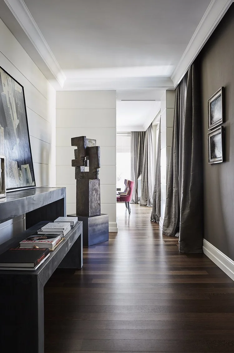 Interno di un'ampia e luminosa stanza con pavimento in legno scuro, pareti bianche e grigie, e tende grigie alle finestre. In primo piano, una console nera con libri e quadri. Al centro, una scultura moderna in pietra. Sullo sfondo, una zona con sedie rosse e grandi finestre che lasciano entrare molta luce.