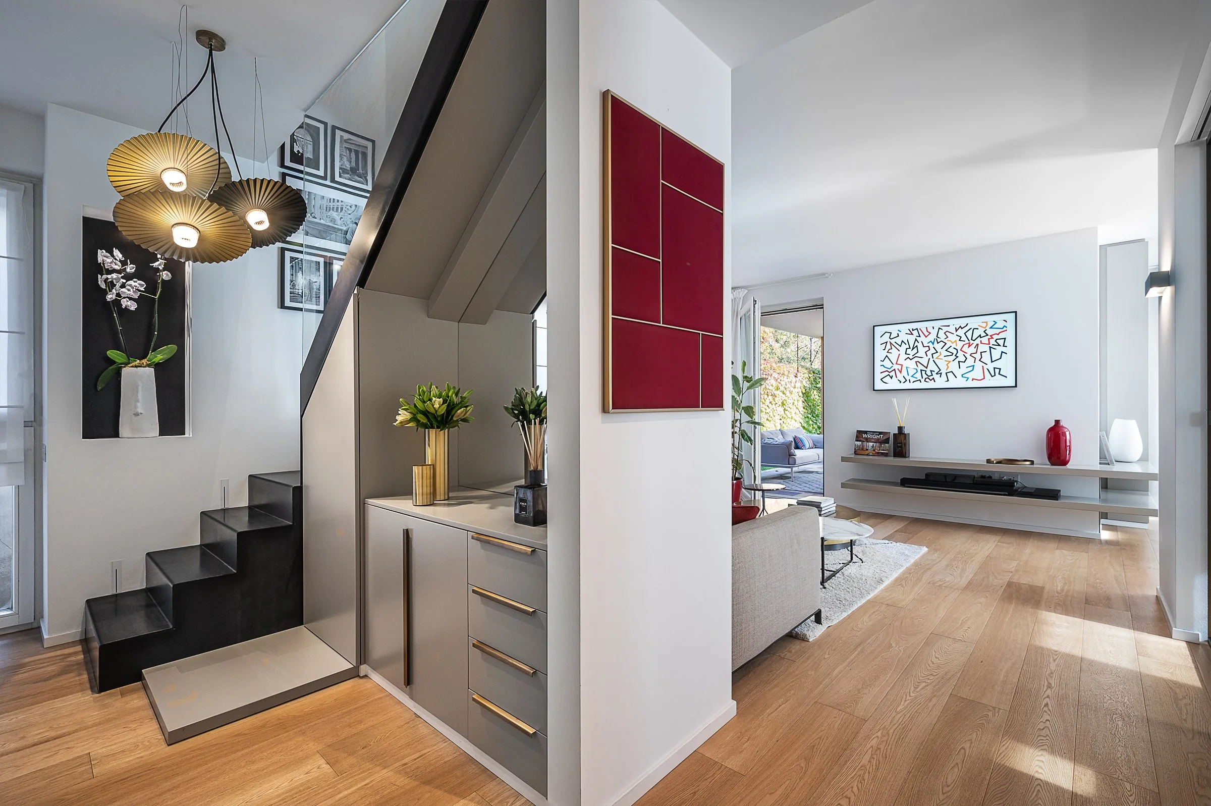 Interno di un appartamento moderno con scala nera, pareti bianche, decorazioni con piante e quadri, vista sul soggiorno con divano, tavolino e mobile TV, pavimento in legno chiaro.