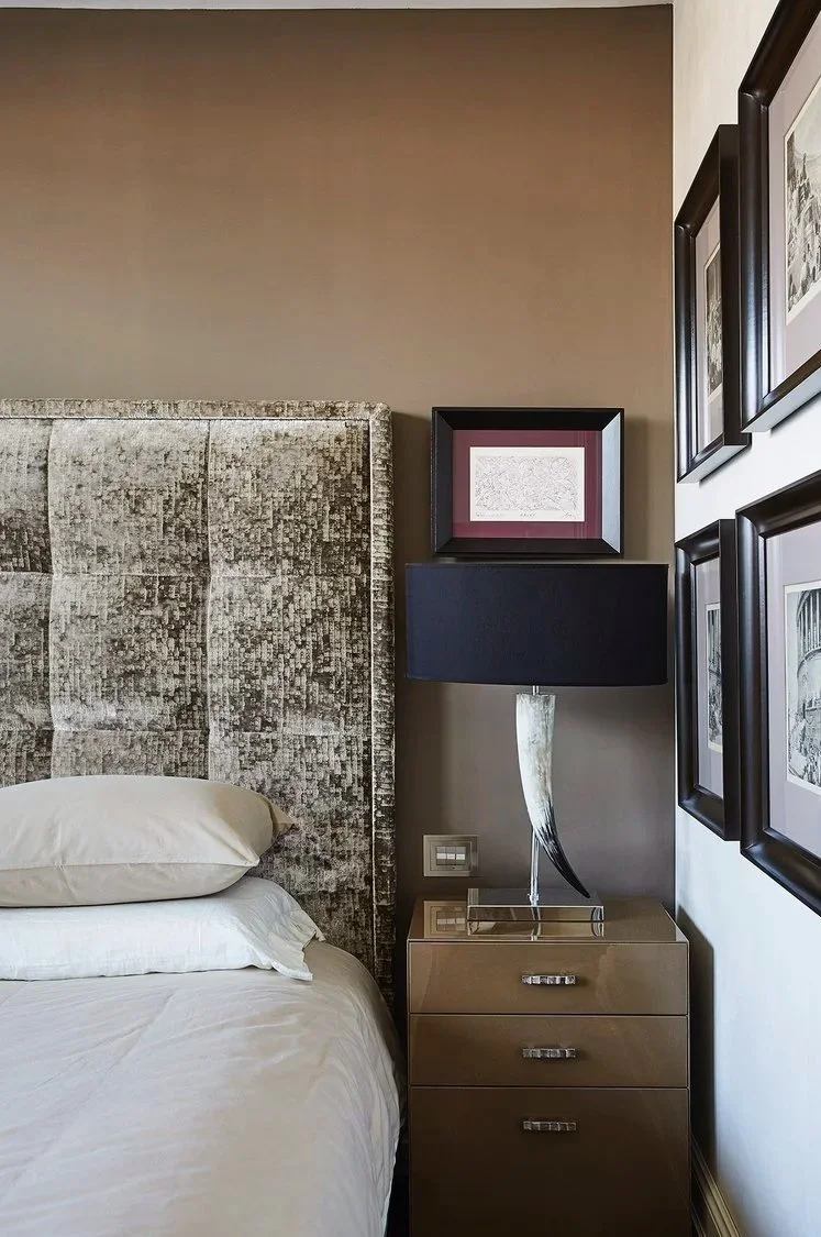 Angolo di una camera da letto con testata imbottita in tessuto grigio, lampada con base chiara e paralume nero, quadro incorniciato sopra la lampada, e una serie di cornici nere con immagini sulla parete di destra.