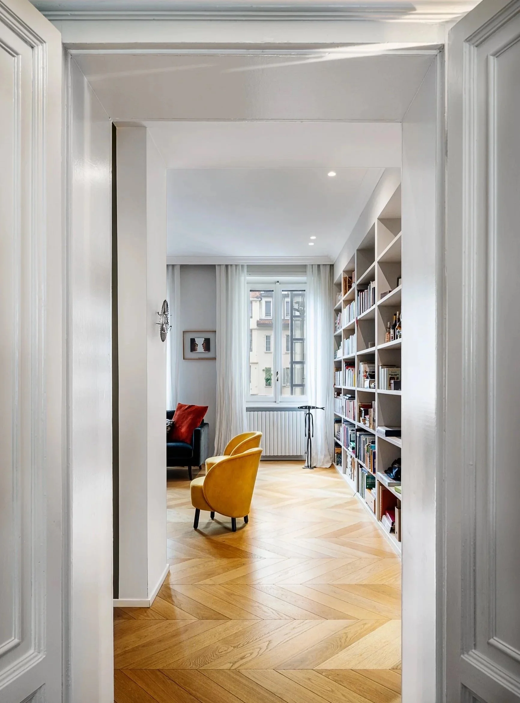 Interno di un soggiorno con libreria bianca, pavimento in parquet, due poltrone gialle e un divano nero con cuscino rosso, finestra grande con tende bianche.