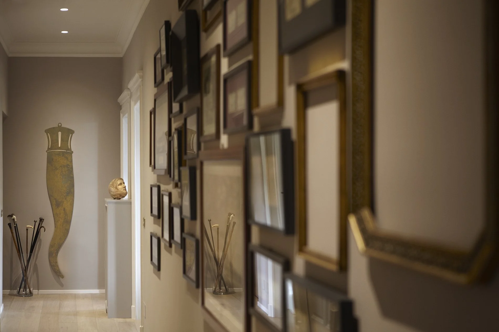 Parete decorata con numerosi quadri, un grande vaso decorativo e una scultura di testa, con luci soffuse e un pavimento in legno.