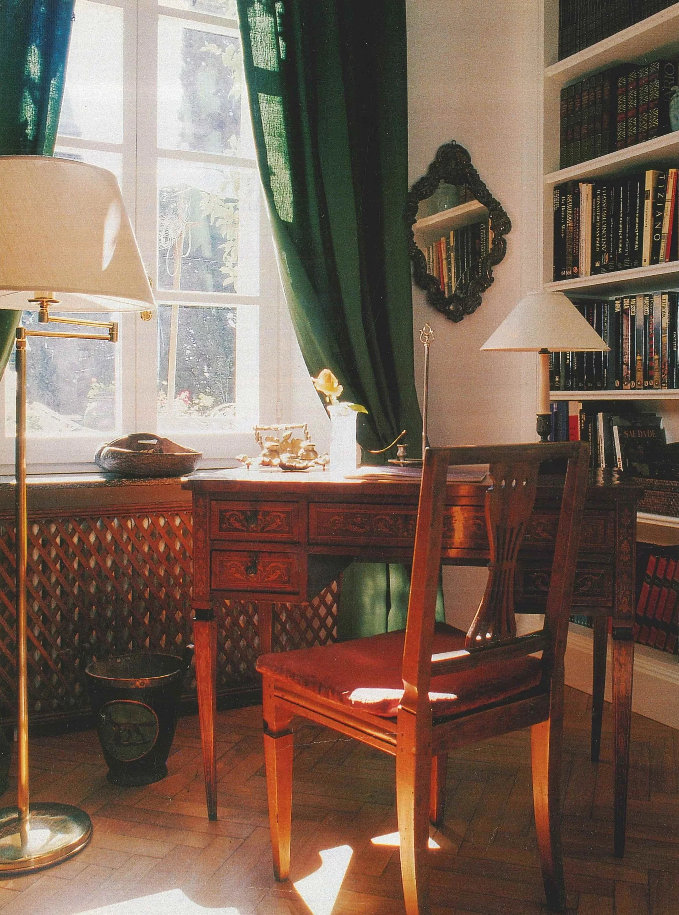 Una stanza con una finestra che entra luce naturale, una scrivania di legno con cassetti, una sedia di legno, scaffali pieni di libri, una lampada da tavolo e decorazioni vari, tra cui un quadro e un vaso con piante.