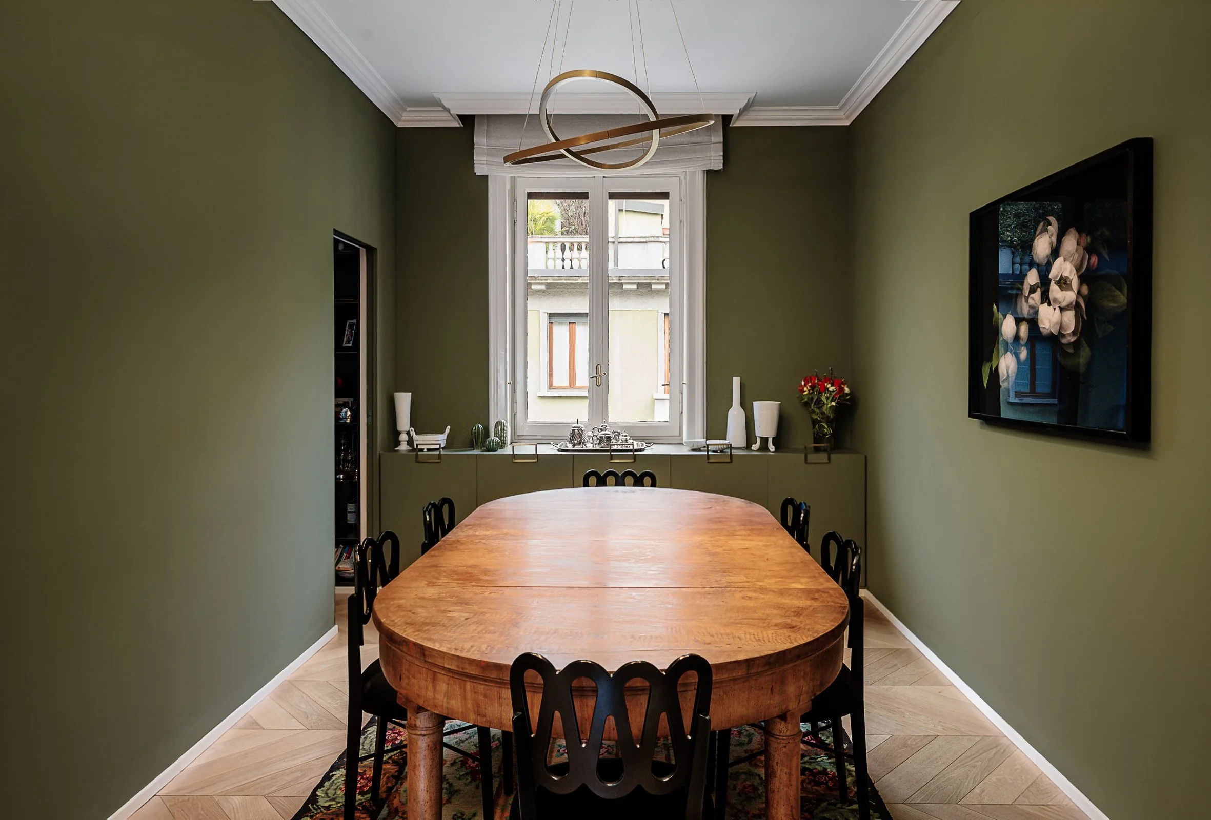 Sala da pranzo con pareti verdi, tavolo in legno e sedie nere, finestra centrale con tende bianche, decorazioni e oggetti sul davanzale, quadro con fiori sulla parete destra, lampadario moderno sopra il tavolo.