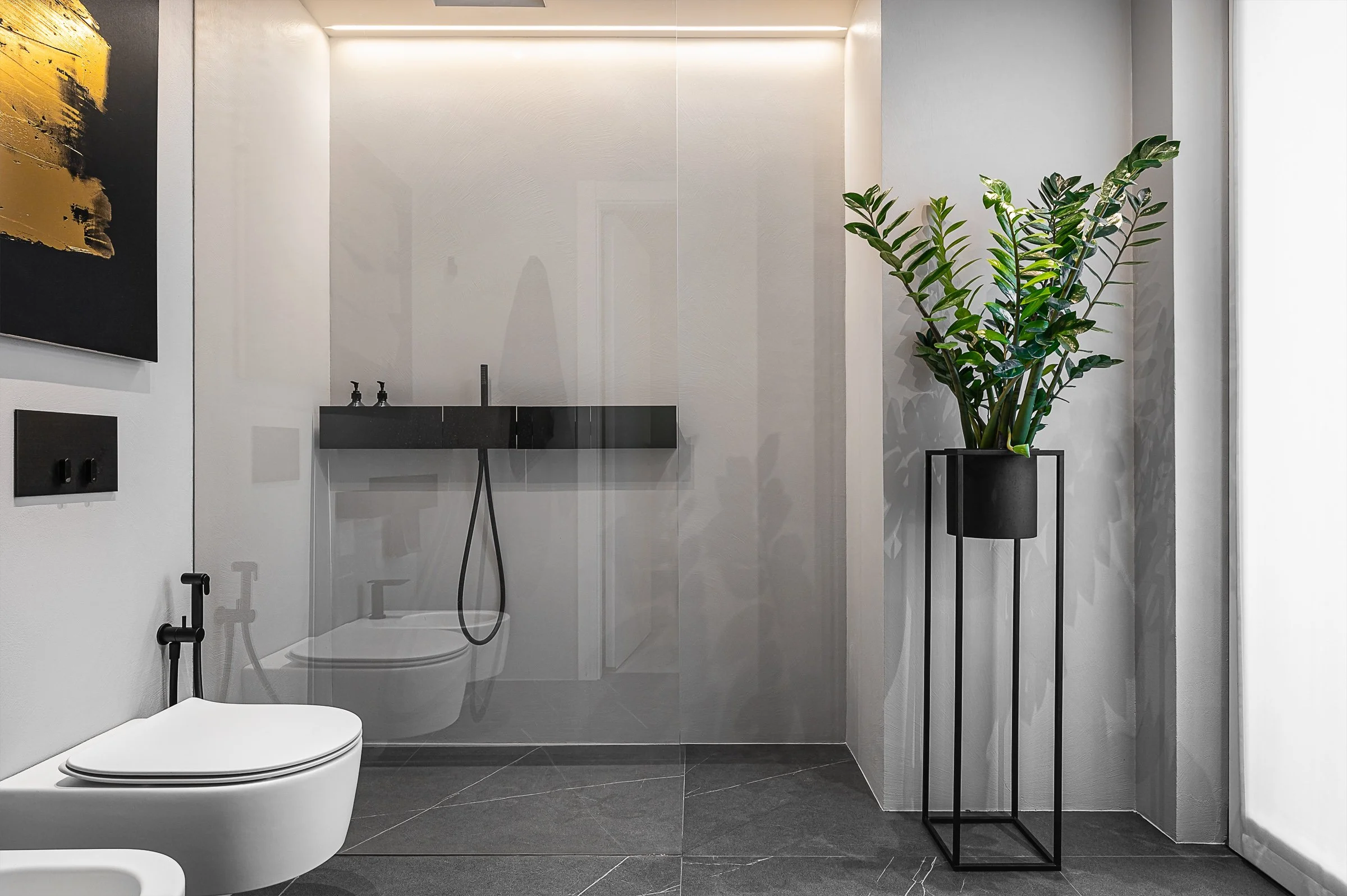 Bagno moderno con pareti bianche, pianta verde in vaso nero su supporto nero, box doccia con porta in vetro, WC bianco.