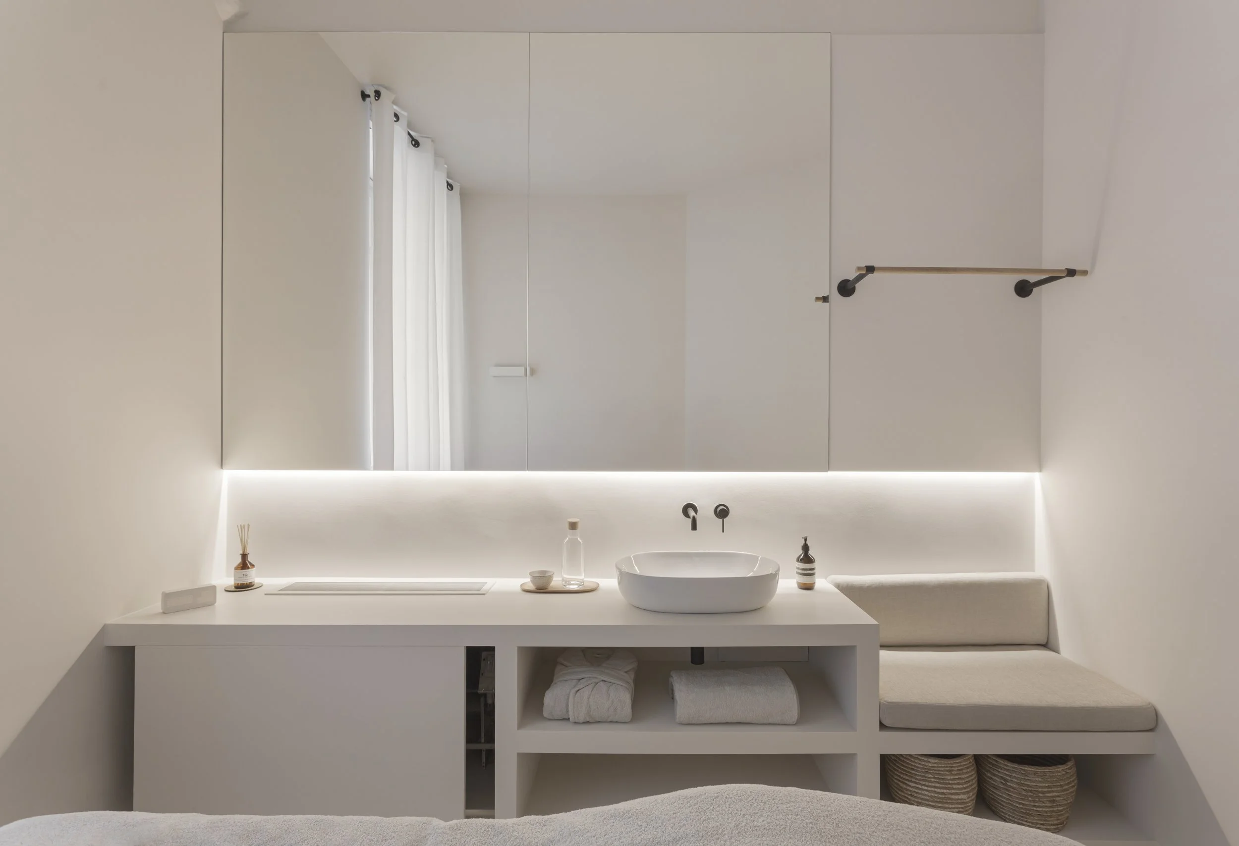 Bagno minimalista con grande specchio, lavabo bianco, sgabello con cuscino, tende bianche, bottiglia di sapone e decorazione con bastoncini diffusori
