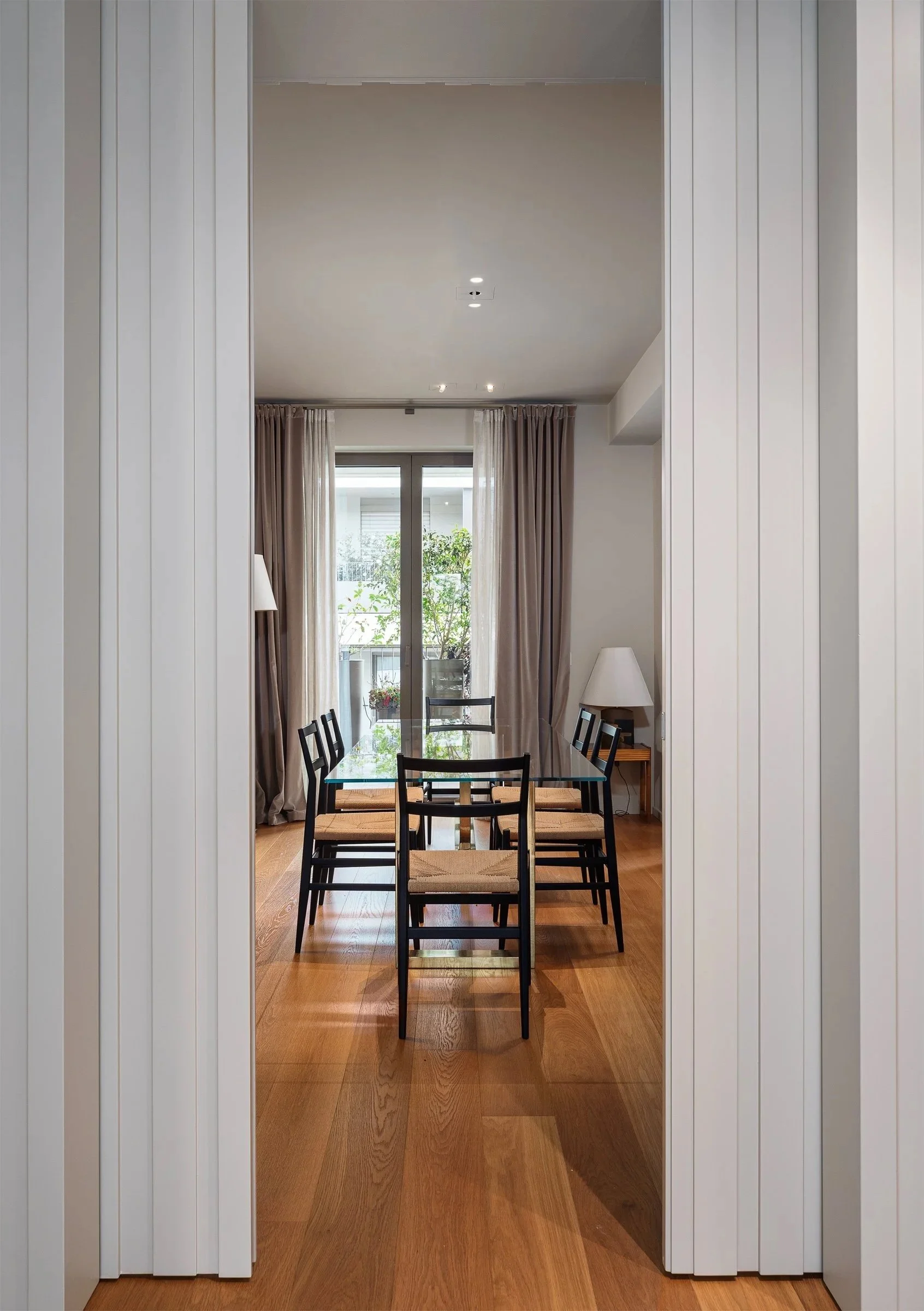 Sala da pranzo con tavolo rettangolare in vetro, sei sedie con struttura nera e sedili in tessuto beige. Sul lato destro, una lampada da tavolo su un tavolino di legno, e una finestra grande con tende beige e una porta finestra che dà su un balcone con piante verdi.