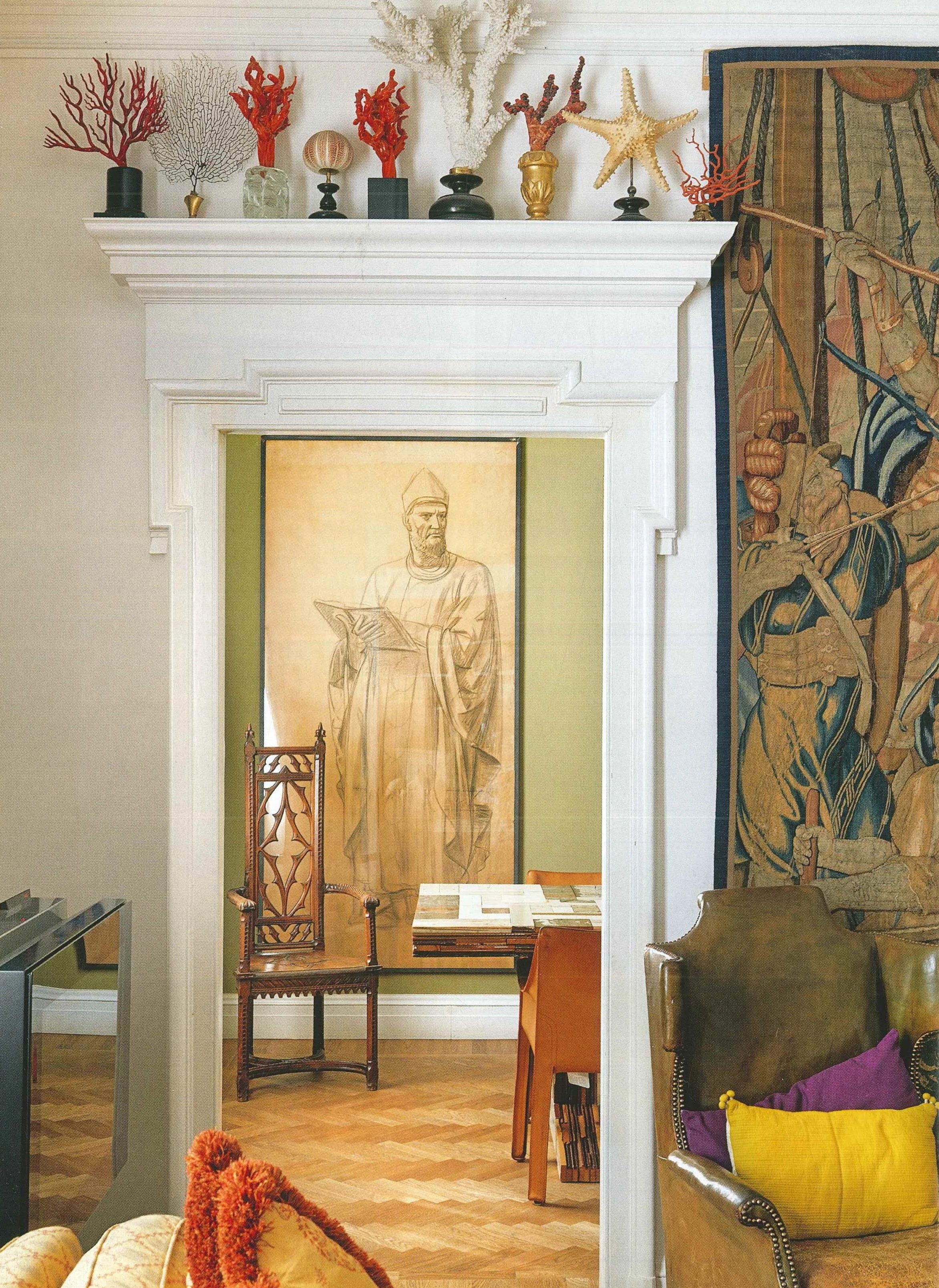 Interno di una stanza con un camino bianco decorato con conchiglie e rami colorati di corallo e stelle marine decorative, un'illustrazione di un uomo con toga e copricapo in uno sfondo verde, mobili in legno e una grande tappeto con un disegno artistico.