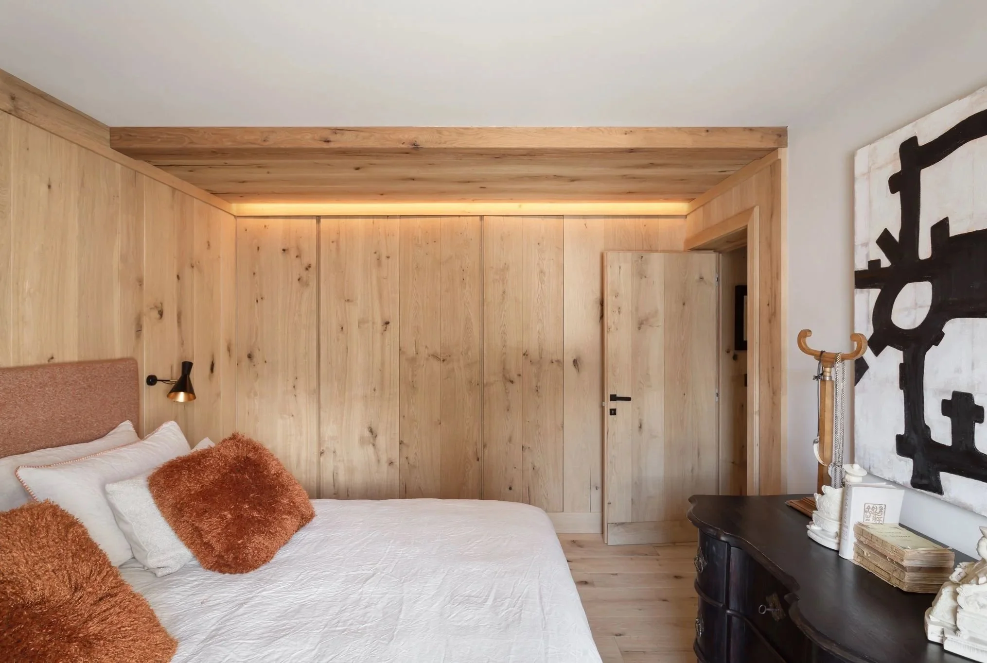 Una camera da letto con pareti e soffitto in legno chiaro, un letto con cuscini marroni e bianchi, una lampada da lettura nera e oro, una cassettiera nera con oggetti decorativi e un grande quadro nero e bianco sulla parete.